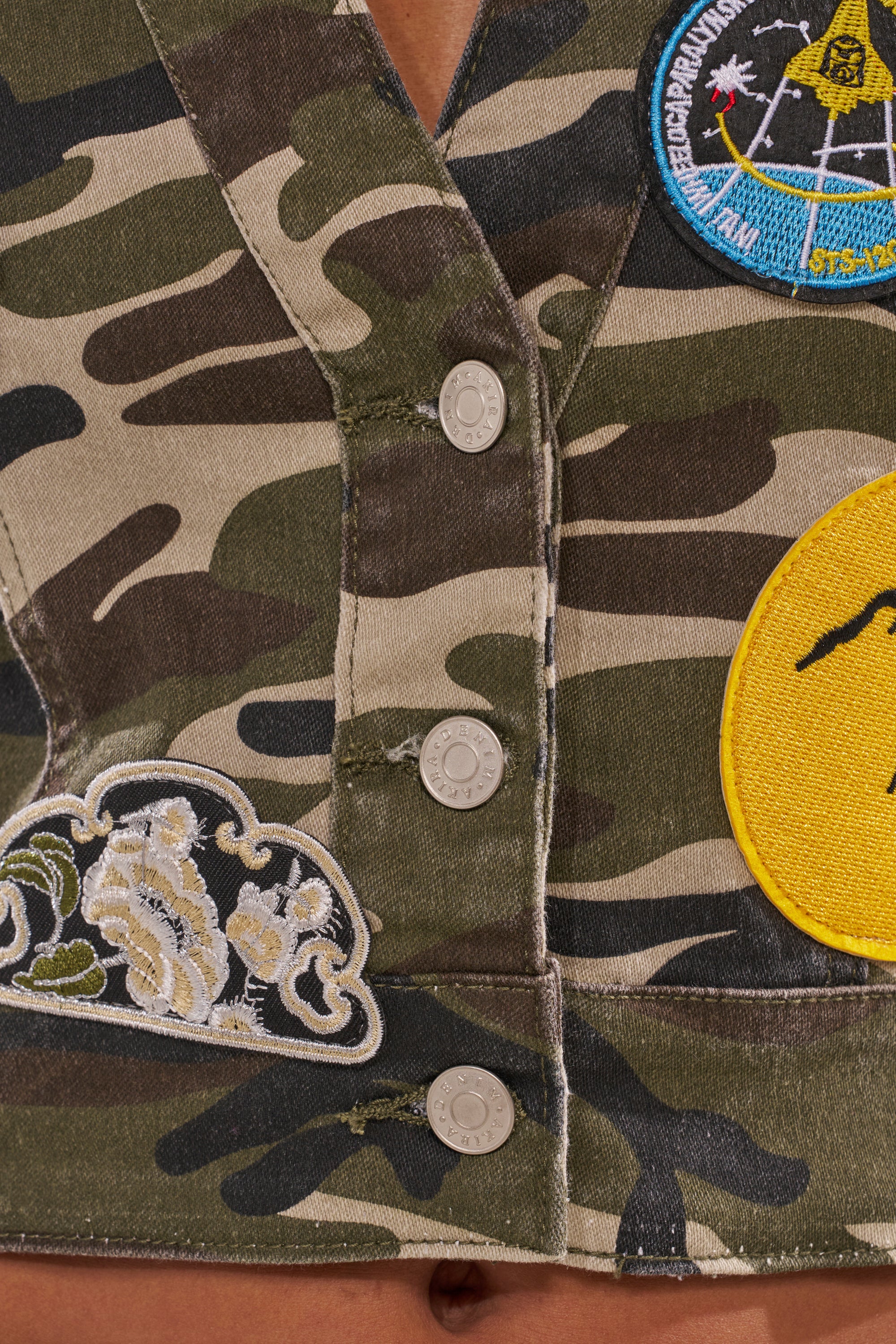 Close-up of the DOWN FOR SOME FUN CAMO BUTTON DOWN vest, featuring silver snap buttons and embroidered patches like a yellow circle, wave, and colorful NASA-themed patch—a bold military-inspired streetwear piece. 8