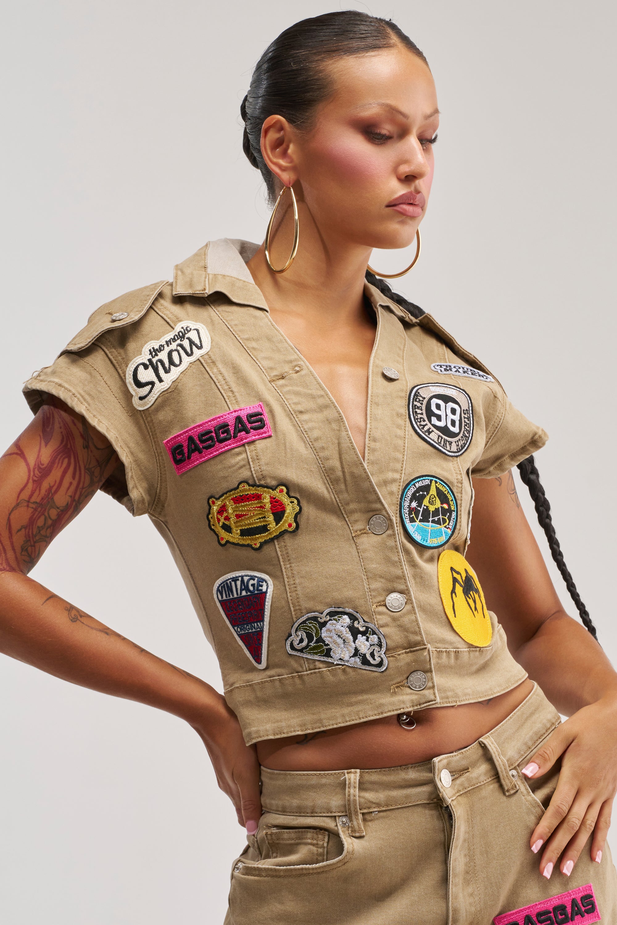 A woman wears the "DOWN FOR SOME FUN BUTTON DOWN," a beige cropped short-sleeve top with matching pants, both featuring colorful embroidered patches such as gas pumps, snowflakes, and mountains. She poses confidently with one hand on her hip.