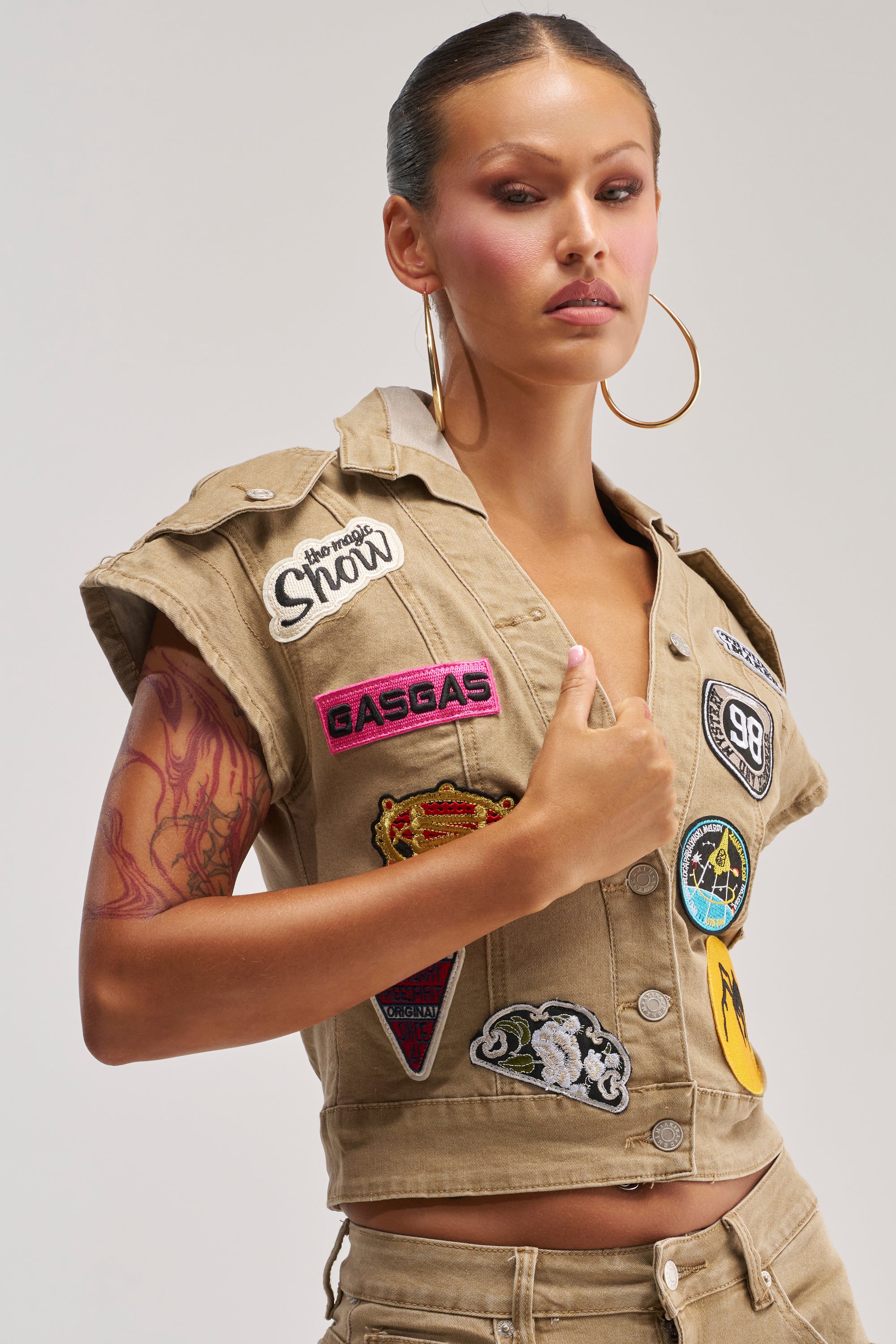 A woman with hoop earrings and pink eyeshadow wears the DOWN FOR SOME FUN BUTTON DOWN—a beige, short-sleeved cropped vest decorated with colorful embroidered patches. She shows off a tattoo on her right arm while posing against a plain light background.