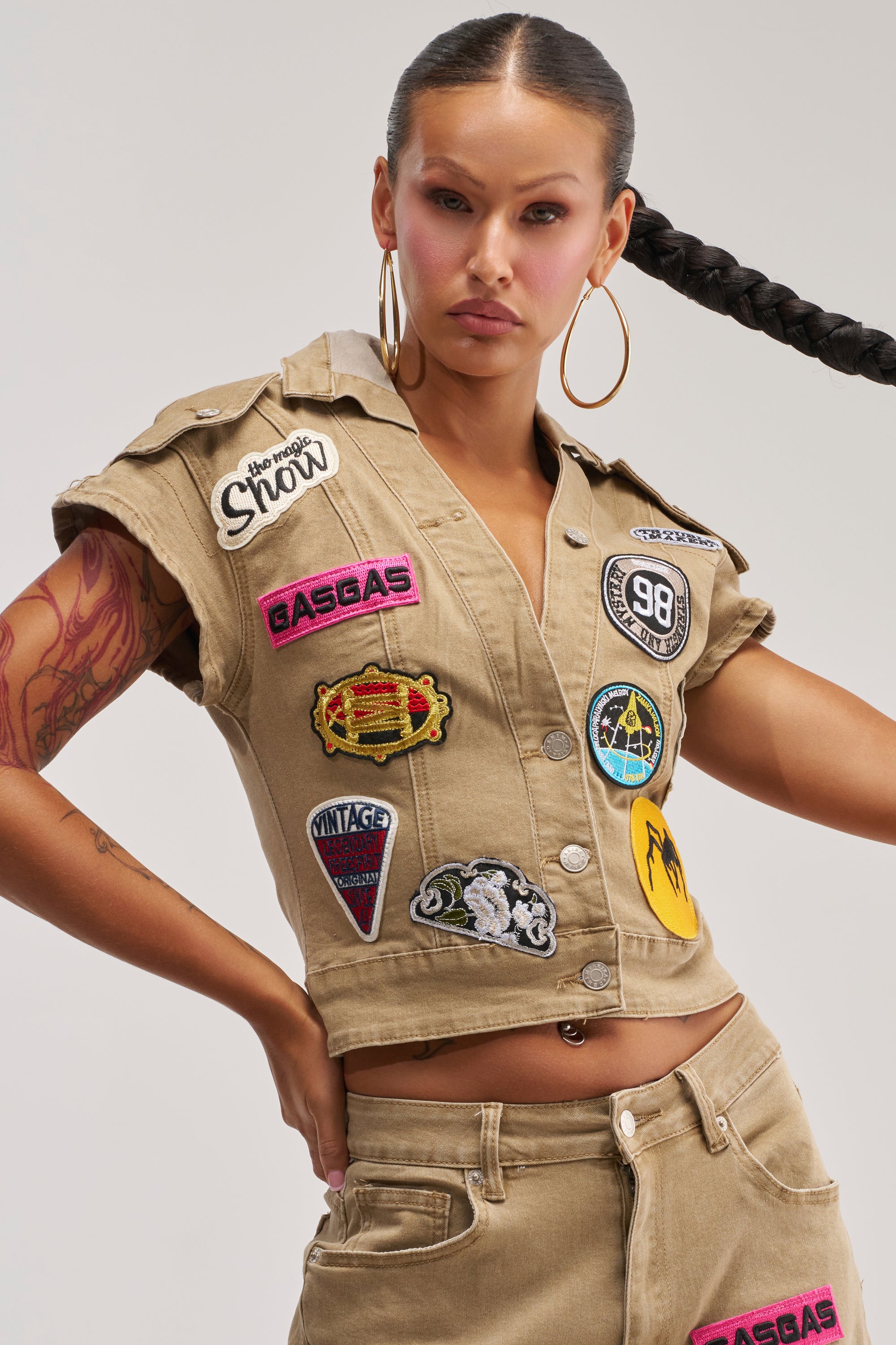 A woman with long braided hair wears the DOWN FOR SOME FUN BUTTON DOWN—a tan, short-sleeve, collared top adorned with colorful embroidered patches—paired with matching pants. She poses confidently with one hand on her hip against a plain background.