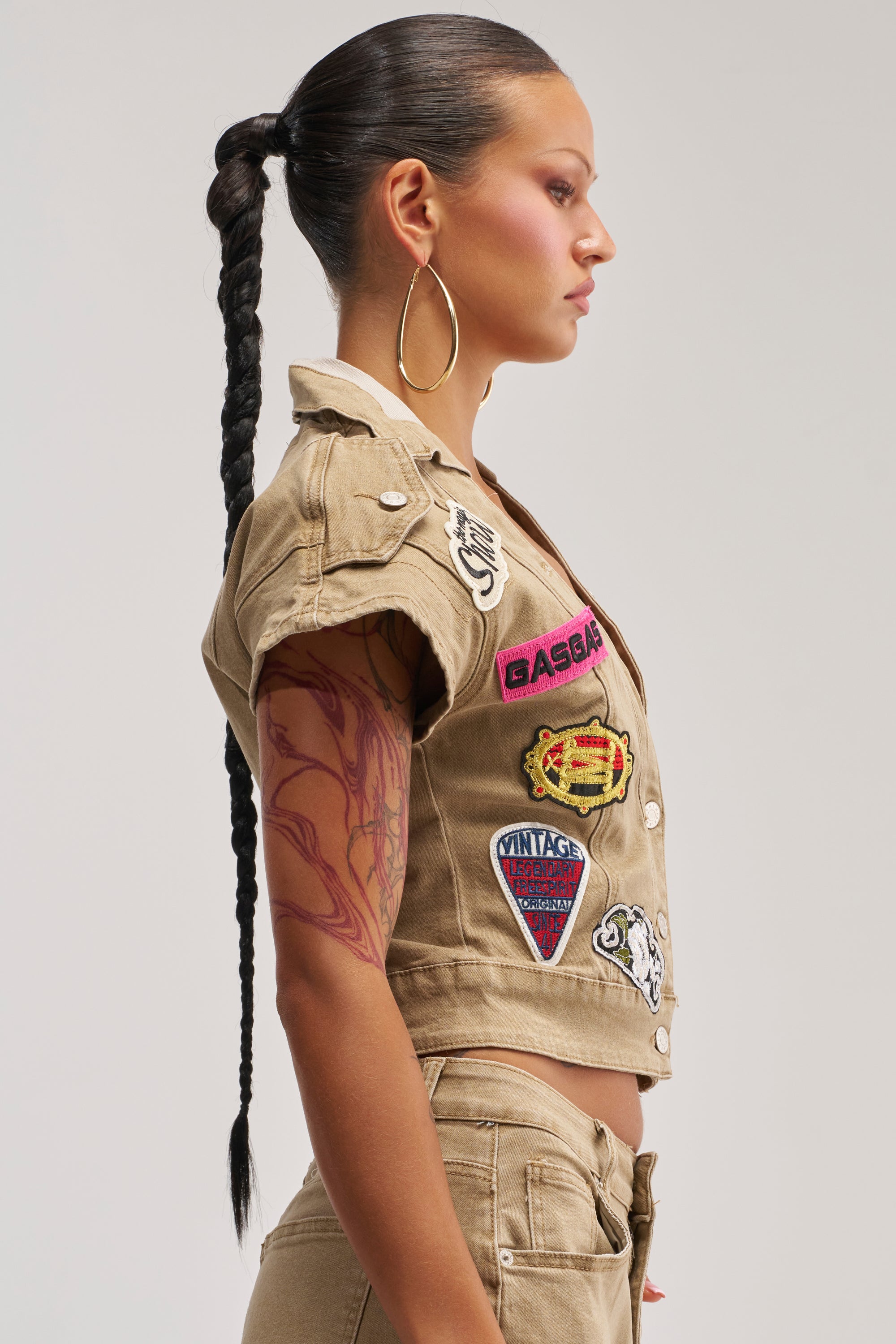 A woman in profile with a long braided ponytail and large hoop earrings wears the DOWN FOR SOME FUN BUTTON DOWN— a beige cropped jacket paired with pants featuring colorful embroidered patches. She displays a red tattoo on her arm against a white background.