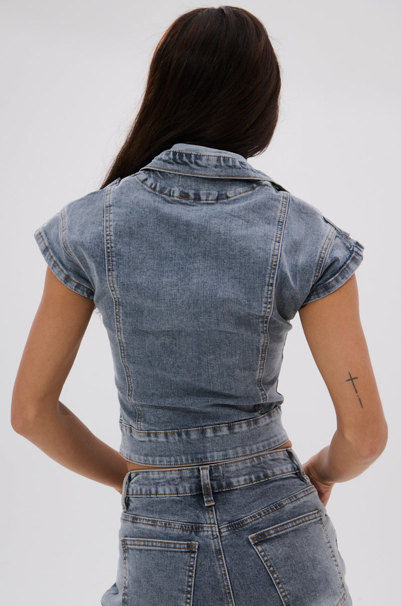 A woman with long dark hair, shown from the back, wears the VINTAGE DENIM BLOUSE WITH PATCHES in a slim fit and high-waisted jeans. A small cross tattoo is on her upper left arm. The background is plain and light. 4