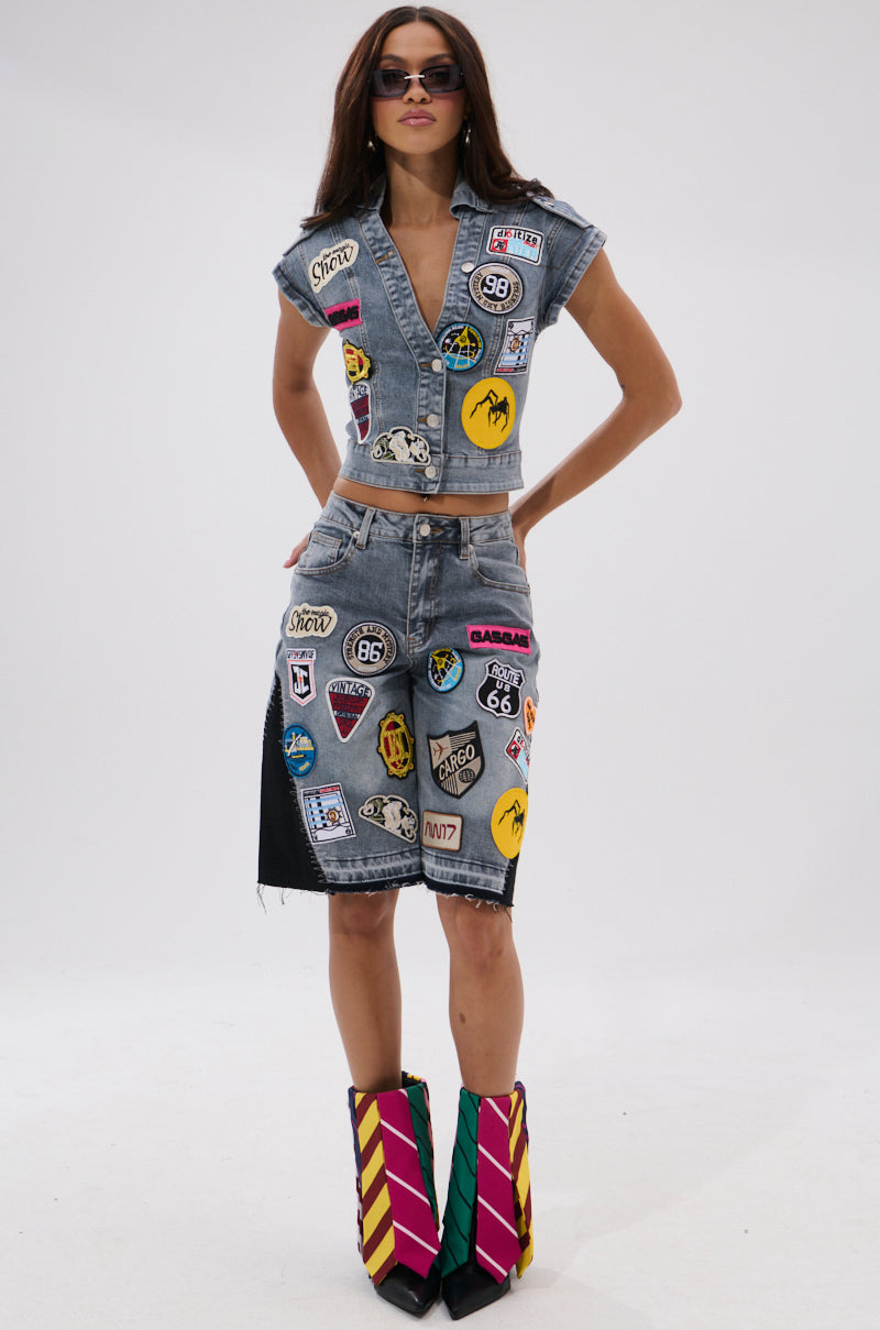 A woman models the VINTAGE DENIM BLOUSE WITH PATCHES and a matching skirt, both sleeveless and cropped with colorful patches. She completes the look with angular sunglasses and geometric multicolored boots against a white background. 5