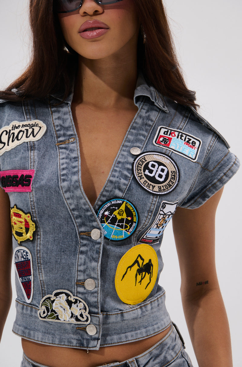 Woman wearing the VINTAGE DENIM BLOUSE WITH PATCHES— a slim-fit denim vest adorned with colorful embroidery, a yellow spider patch, “98” badge, and other unique designs—paired with stylish sunglasses. 7