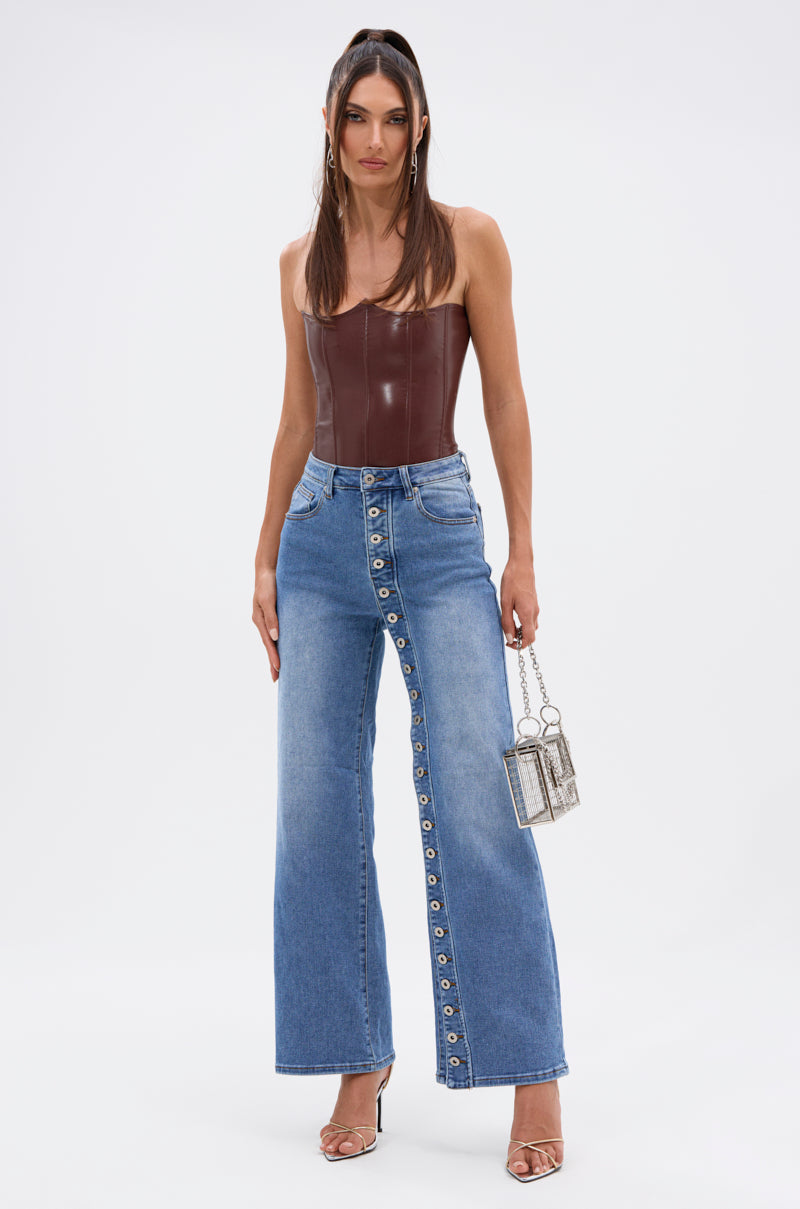 A woman stands against a white background wearing the SNAP OUT OF IT DENIM PANT with a straight-leg fit and side button detailing, styled with a brown strapless leather corset, heeled sandals, and a small metallic handbag. 0