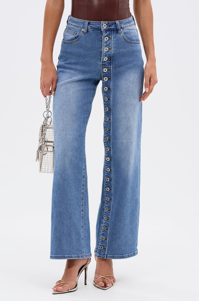 A woman wearing the SNAP OUT OF IT DENIM PANT, featuring a wide-leg, mid-rise design with front button closure. She holds a small silver handbag and wears silver strappy heels; upper body and face are not shown. 1