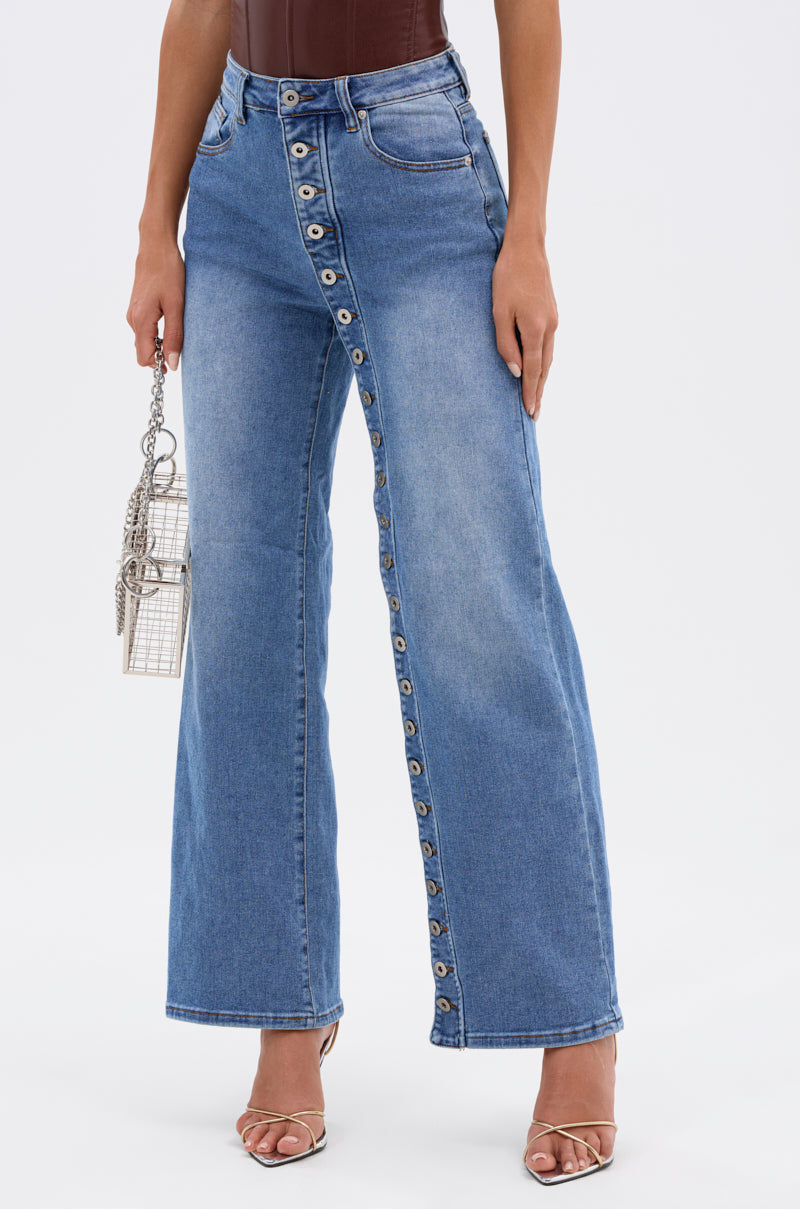 A person wearing the SNAP OUT OF IT DENIM PANT—high-waisted, straight-leg blue denim with a diagonal button closure—paired with a small metallic handbag and open-toed heels. 2