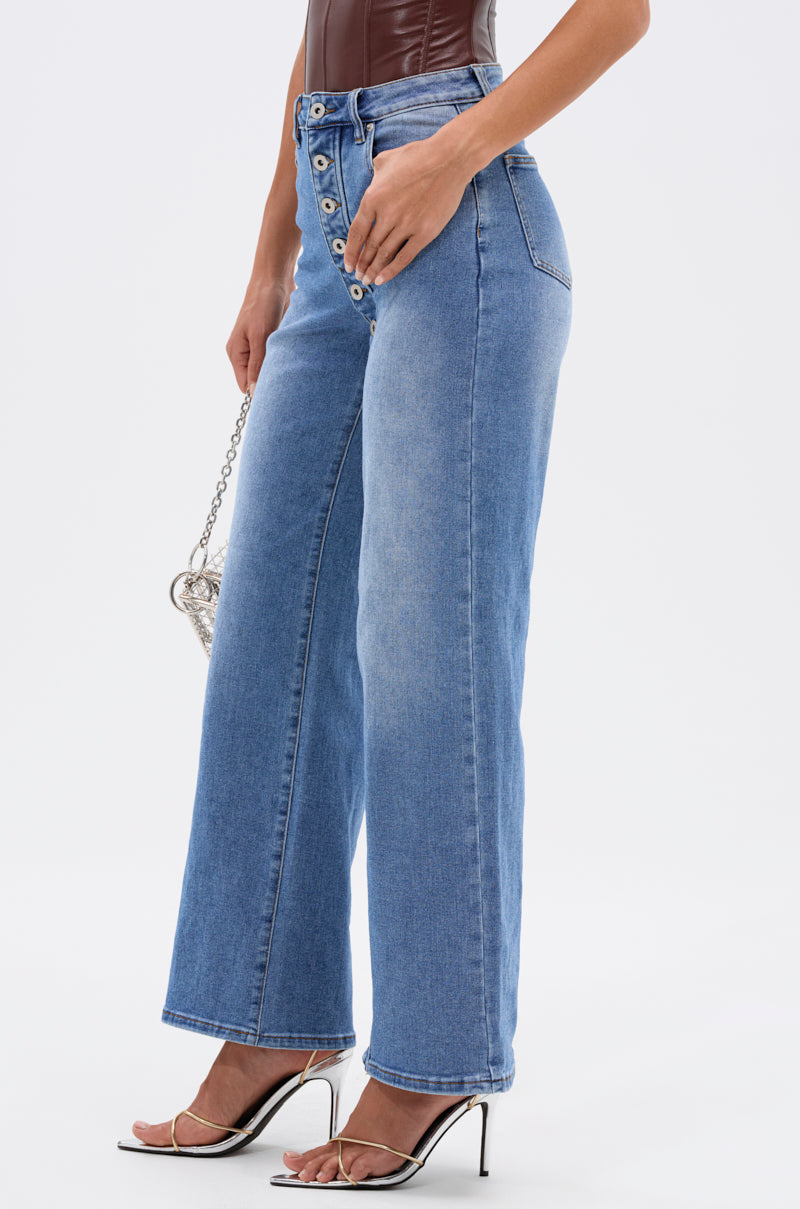 A person wears the SNAP OUT OF IT DENIM PANT with front buttons, a brown strapless top, and white heeled sandals, holding a small silver chain bag against a plain white background. 3