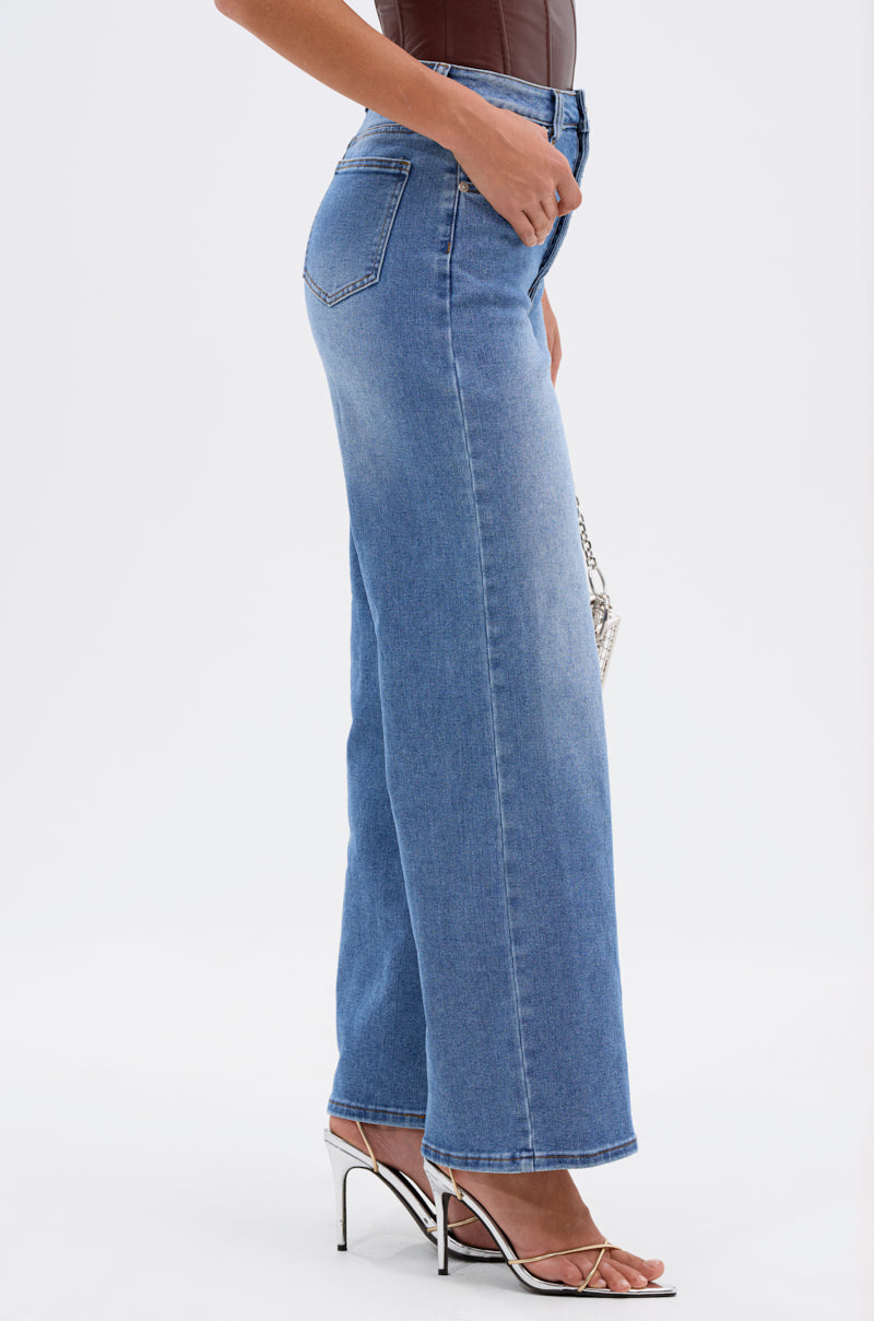 A person wearing the SNAP OUT OF IT DENIM PANT in blue with white high-heeled sandals, shown from the side and holding a small silver handbag against a plain white background for a timeless, chic look. 4