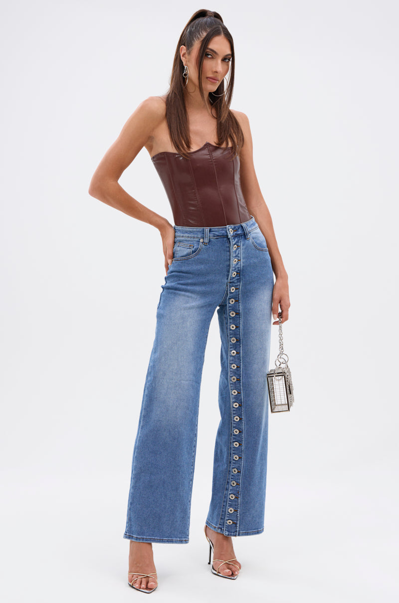 A woman poses in the SNAP OUT OF IT DENIM PANT, featuring mid-rise fit and diagonal button details, styled with a maroon strapless leather top, clear heeled sandals, and a small metallic handbag against a white background. 6