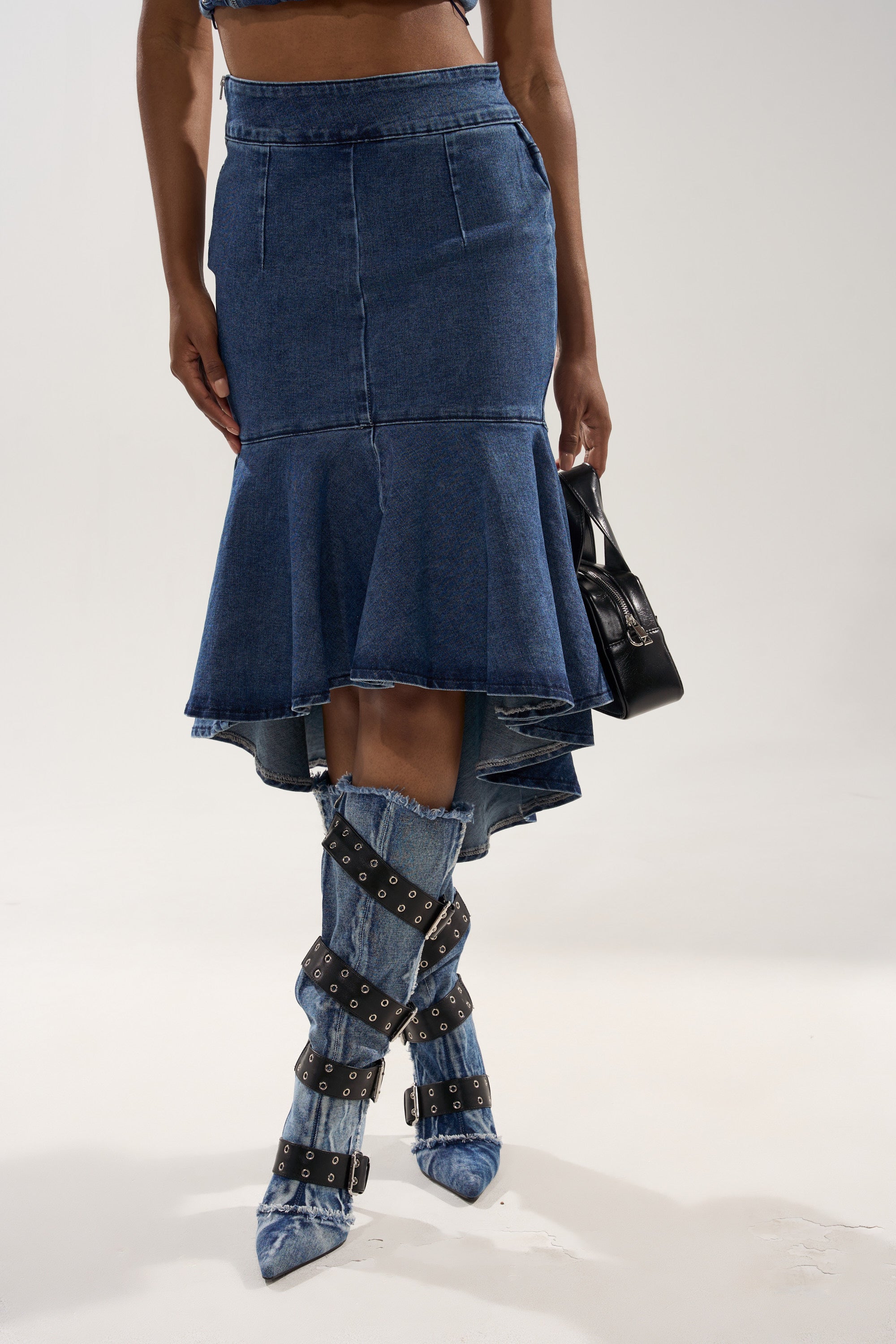 A person wears the YOU WISH DENIM MIDI SKIRT, a body-skimming, high-low denim midi skirt with an asymmetrical ruffled hem, styled with knee-high denim boots, black straps and buckles, and a small black handbag against a plain background. 0