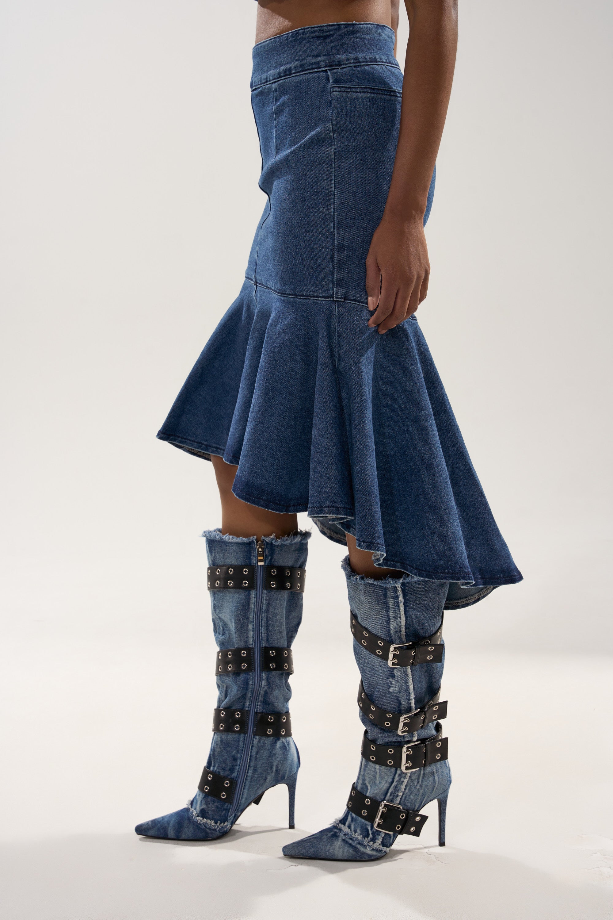 A person wearing the YOU WISH DENIM MIDI SKIRT, a contoured blue denim midi skirt with an asymmetrical ruffled hem, styled with matching knee-high denim boots featuring multiple black buckle straps, stands against a plain light background. 2
