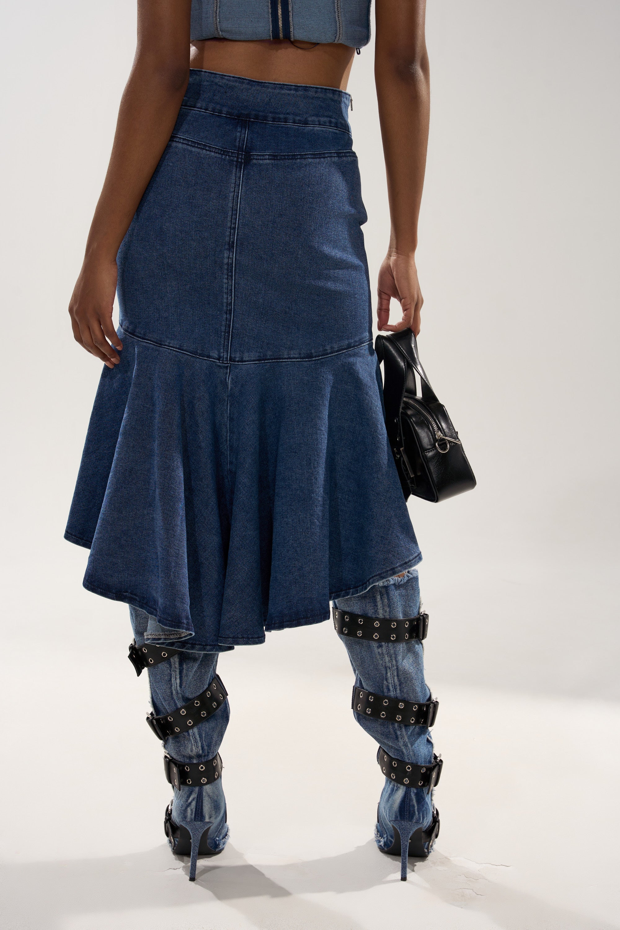 Someone wearing the YOU WISH DENIM MIDI SKIRT over matching denim pants with black strappy buckles, holding a small black handbag. The photo is taken from behind against a plain background. 3