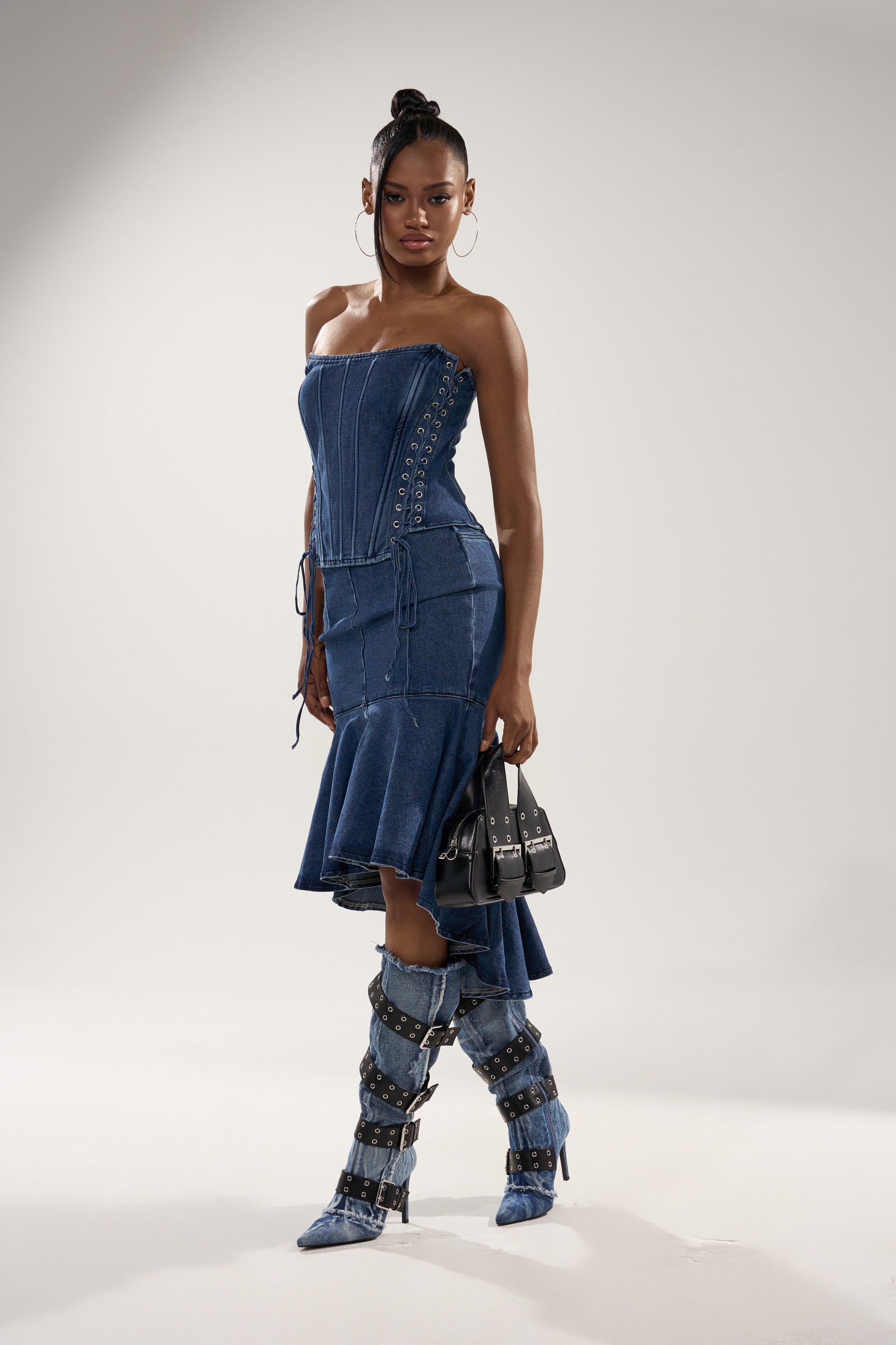 A woman models the YOU WISH DENIM MIDI SKIRT, featuring a contoured fit, asymmetrical ruffled hem, and lace-up details. She pairs it with matching strappy knee-high boots, a small black handbag, and hoop earrings against a light gray background. 4