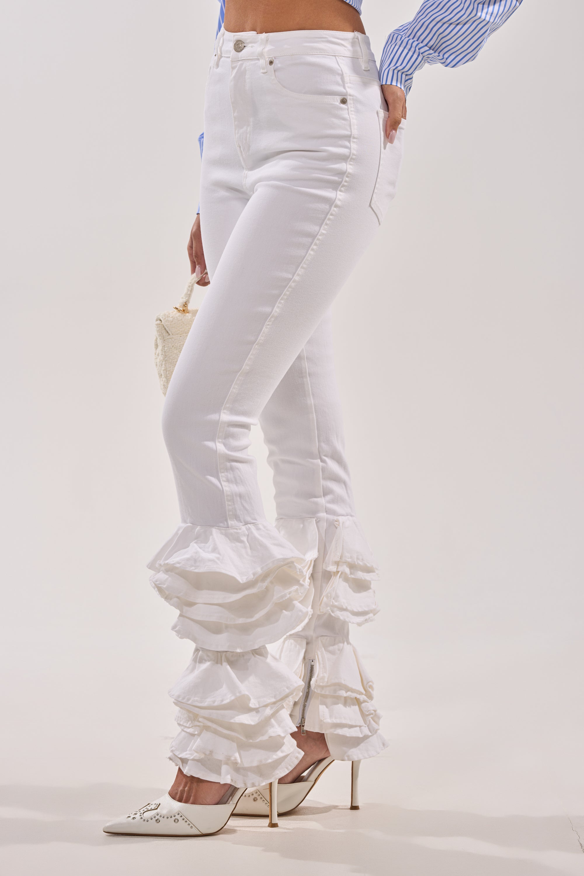 A person wears the NOT ENOUGH RUFFLES DENIM high-rise skinny jeans with layered ankle ruffles, a blue and white striped blouse, white heels, and carries a small textured handbag against a plain background. 2