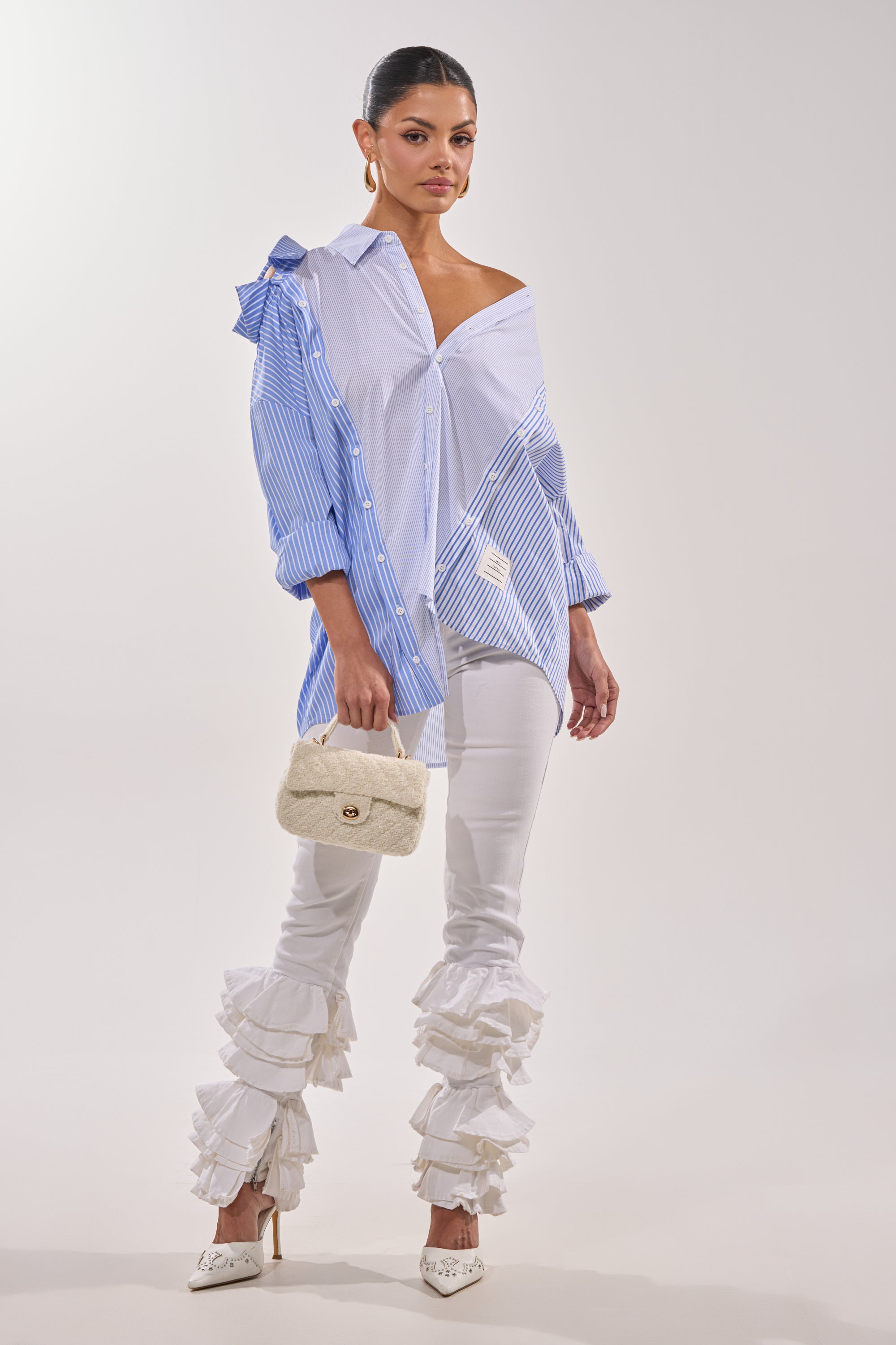 A woman stands against a plain light background wearing NOT ENOUGH RUFFLES DENIM jeans with ruffled hems, a blue and white striped asymmetrical shirt, white heels, and holding a small quilted white bag. 5