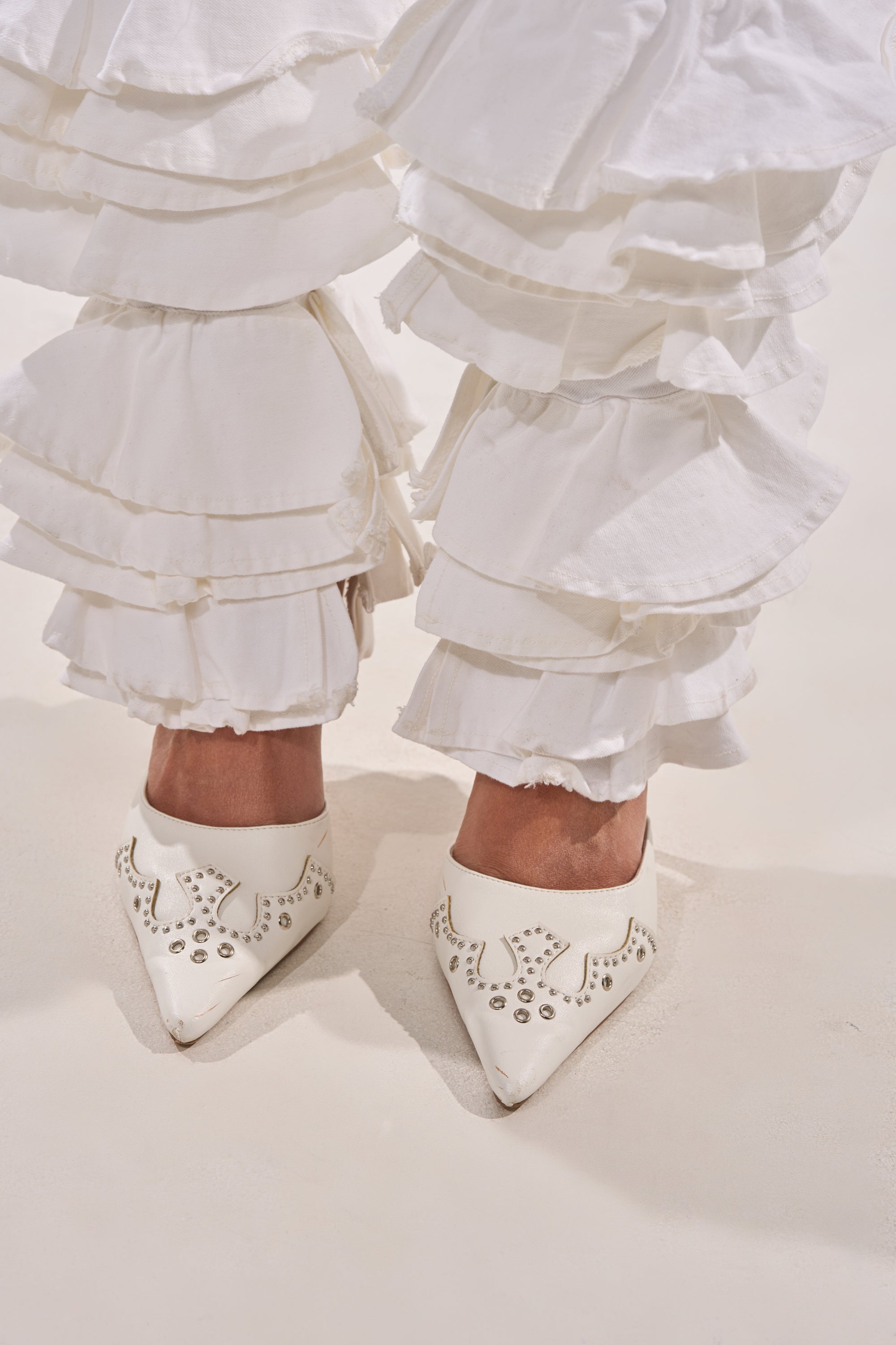 A person stands on a light surface wearing NOT ENOUGH RUFFLES DENIM pants paired with pointed white shoes featuring decorative cut-out patterns. 7