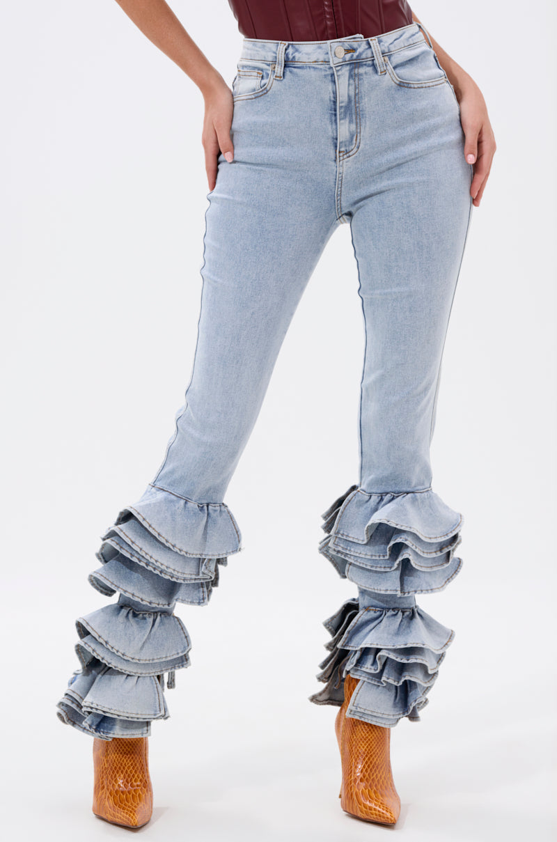 A person models the NOT ENOUGH RUFFLE DENIM high-rise skinny jeans with a multi-tiered ruffle hem, paired with a burgundy top and tan heeled boots against a plain white background.