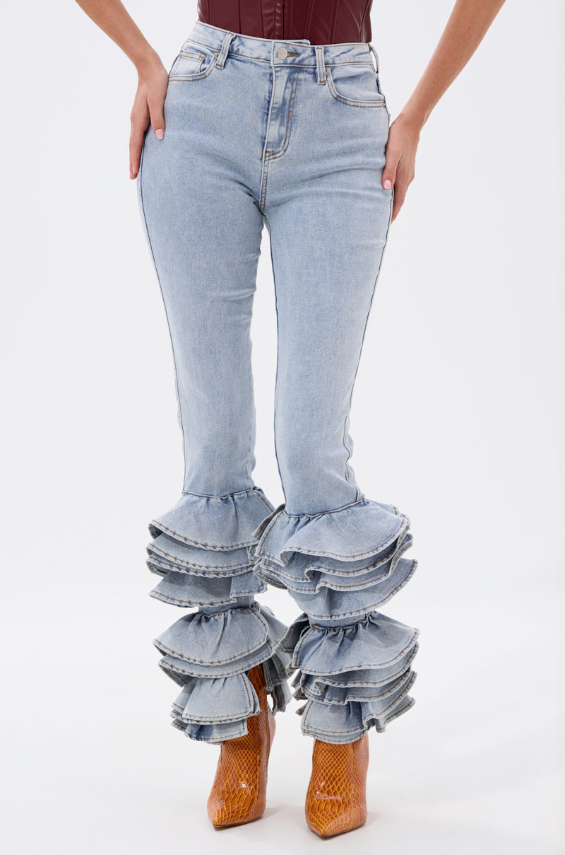 A woman stands against a plain white background wearing the NOT ENOUGH RUFFLE DENIM high-rise skinny jeans with dramatic ruffle hems and brown textured ankle boots.