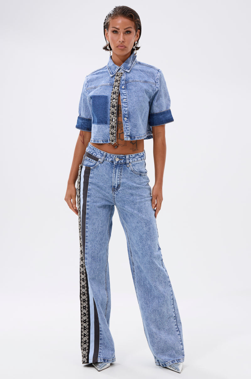 A woman faces forward in the ELEVATED RELAXED DENIM jacket and matching high-waisted, wide-leg jeans with a black-and-white striped detail, her slicked-back hair and confident pose set against a plain background. 0