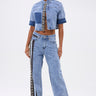 A woman faces forward in the ELEVATED RELAXED DENIM jacket and matching high-waisted, wide-leg jeans with a black-and-white striped detail, her slicked-back hair and confident pose set against a plain background.