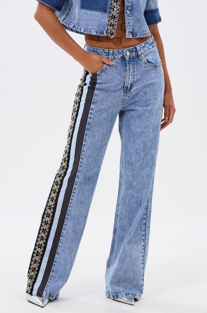 A person models ELEVATED RELAXED DENIM: high-waisted, wide-leg blue jeans with black and white floral side stripes, styled with a cropped denim jacket for a chic, modern look. 1
