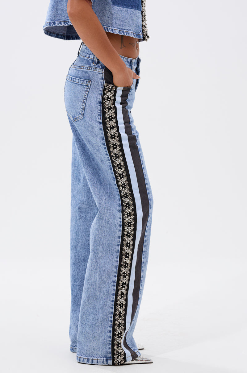 A person wears ELEVATED RELAXED DENIM—light blue, high-waisted, wide-leg jeans with black and white side stripes, floral patterns, and metallic beading—paired with a matching denim crop top, standing against a plain white background. 4