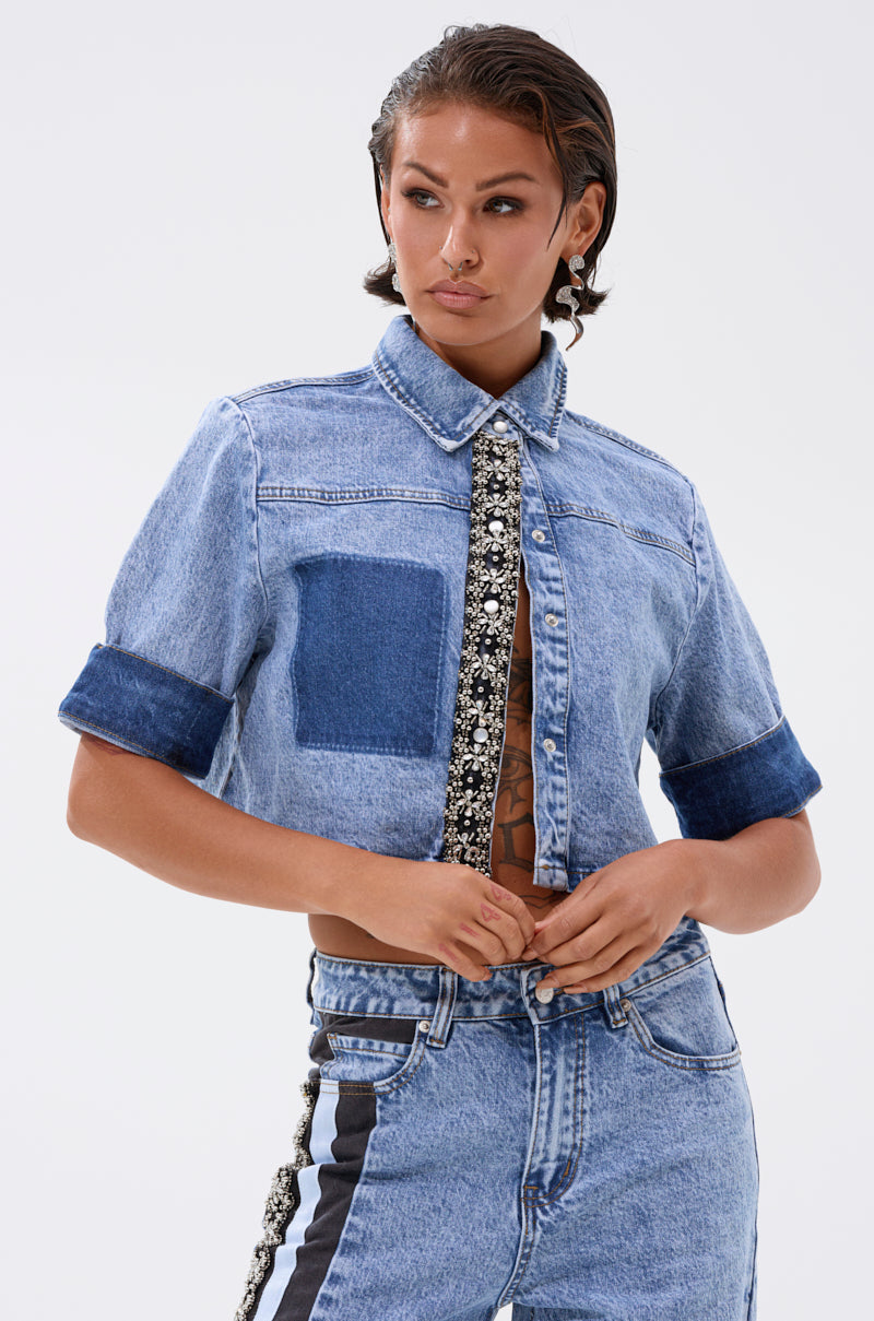 Someone is wearing the ELEVATED DENIM BUTTON DOWN with embellishments, short sleeves, dark cuffs, a patch detail, and decorative trim on the buttons, paired with matching high-waisted jeans featuring a black and white side stripe. 0