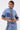 Someone is wearing the ELEVATED DENIM BUTTON DOWN with embellishments, short sleeves, dark cuffs, a patch detail, and decorative trim on the buttons, paired with matching high-waisted jeans featuring a black and white side stripe.
