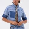 Someone is wearing the ELEVATED DENIM BUTTON DOWN with embellishments, short sleeves, dark cuffs, a patch detail, and decorative trim on the buttons, paired with matching high-waisted jeans featuring a black and white side stripe.