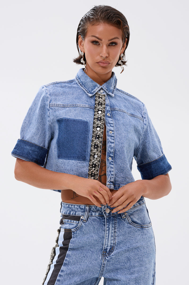 A woman with slicked-back hair wears the ELEVATED DENIM BUTTON DOWN, styled with matching dual-tone, high-waisted jeans featuring black side stripes, standing confidently against a plain background. 1
