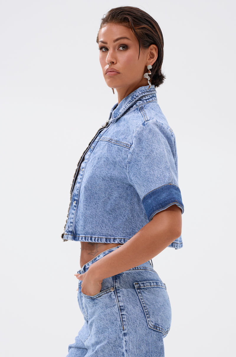A woman stands in profile against a white background, confidently looking at the camera while wearing the ELEVATED DENIM BUTTON DOWN and jeans, one hand casually placed in her pocket. 2