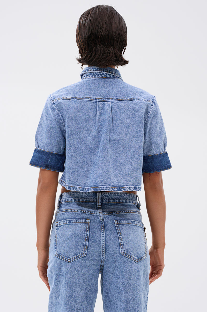 A person with short, dark, wet-look hair wears the ELEVATED DENIM BUTTON DOWN—dual-tone, cropped with rolled sleeves—and high-waisted blue jeans, seen from the back against a plain light background. 3
