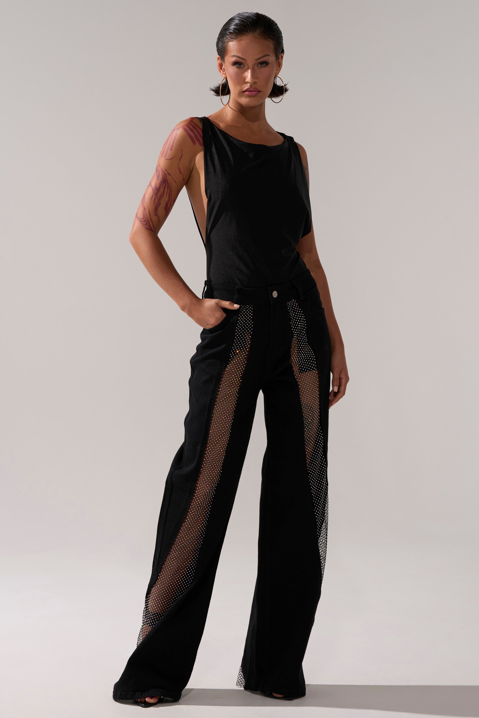 A model poses confidently in the SOMETHING DIFFERENT RELAXED DENIM WITH RHINESTONE MESH, showcasing wide-leg jeans with rhinestone mesh panels down the front, paired with a sleeveless black top against a plain light background.