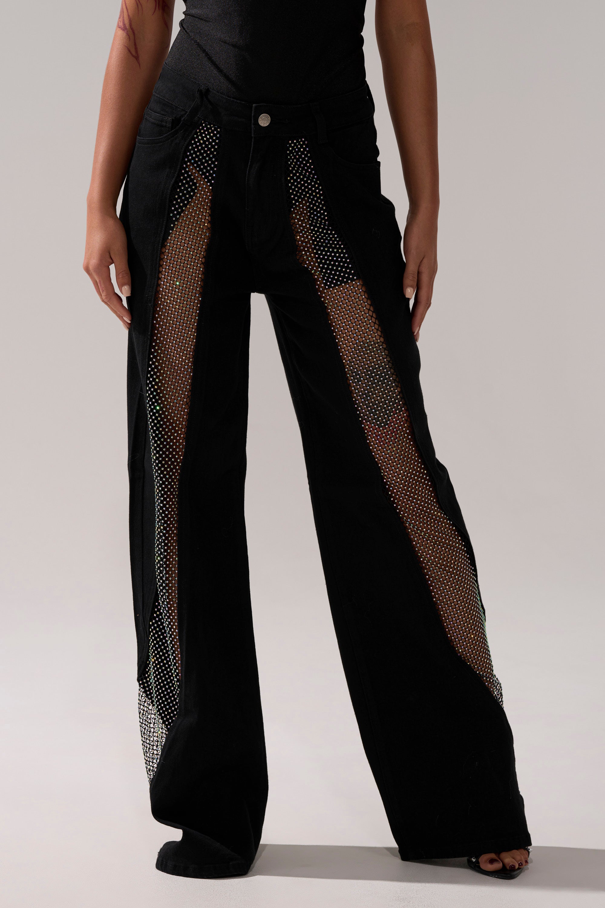 Someone wears the SOMETHING DIFFERENT RELAXED DENIM WITH RHINESTONE MESH—black wide-leg jeans featuring large, curved rhinestone mesh cutouts down the front that reveal skin. The pants have a button closure and are styled with a black top.