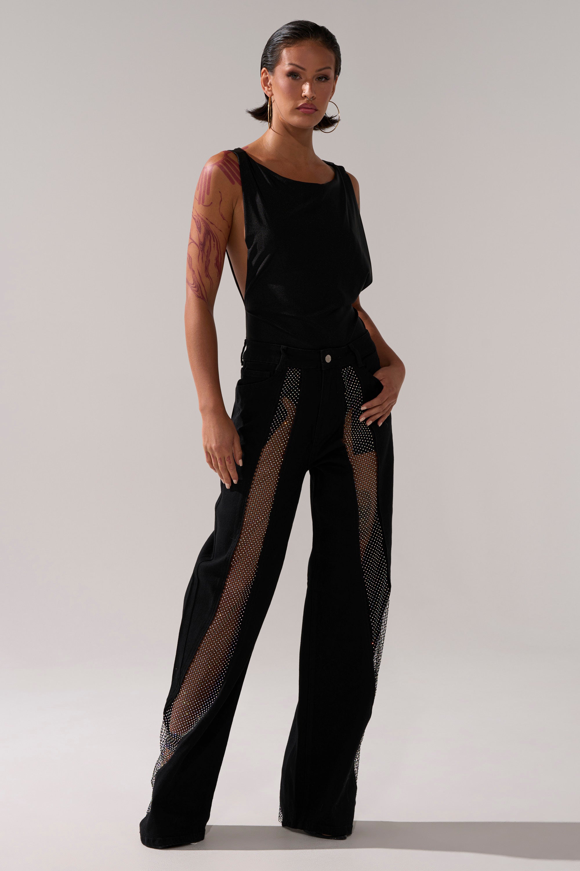 A woman with slicked-back hair stands confidently against a plain, light background, wearing SOMETHING DIFFERENT RELAXED DENIM WITH RHINESTONE MESH and a sleeveless black top, one hand casually in her pocket.
