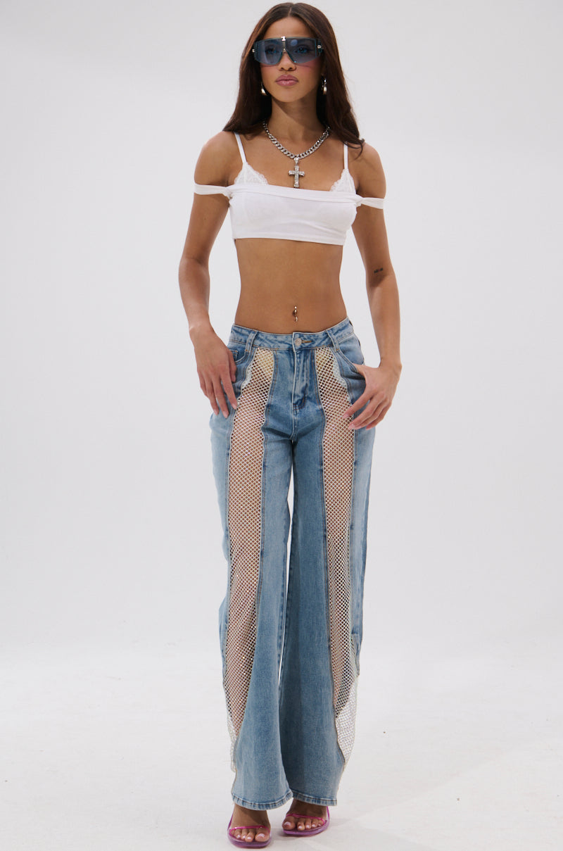 A woman poses against a white background wearing BLING BLING RELAXED DENIM high-rise jeans with mesh panels, a white off-shoulder crop top, pink heels, layered necklaces, and blue sunglasses. 1