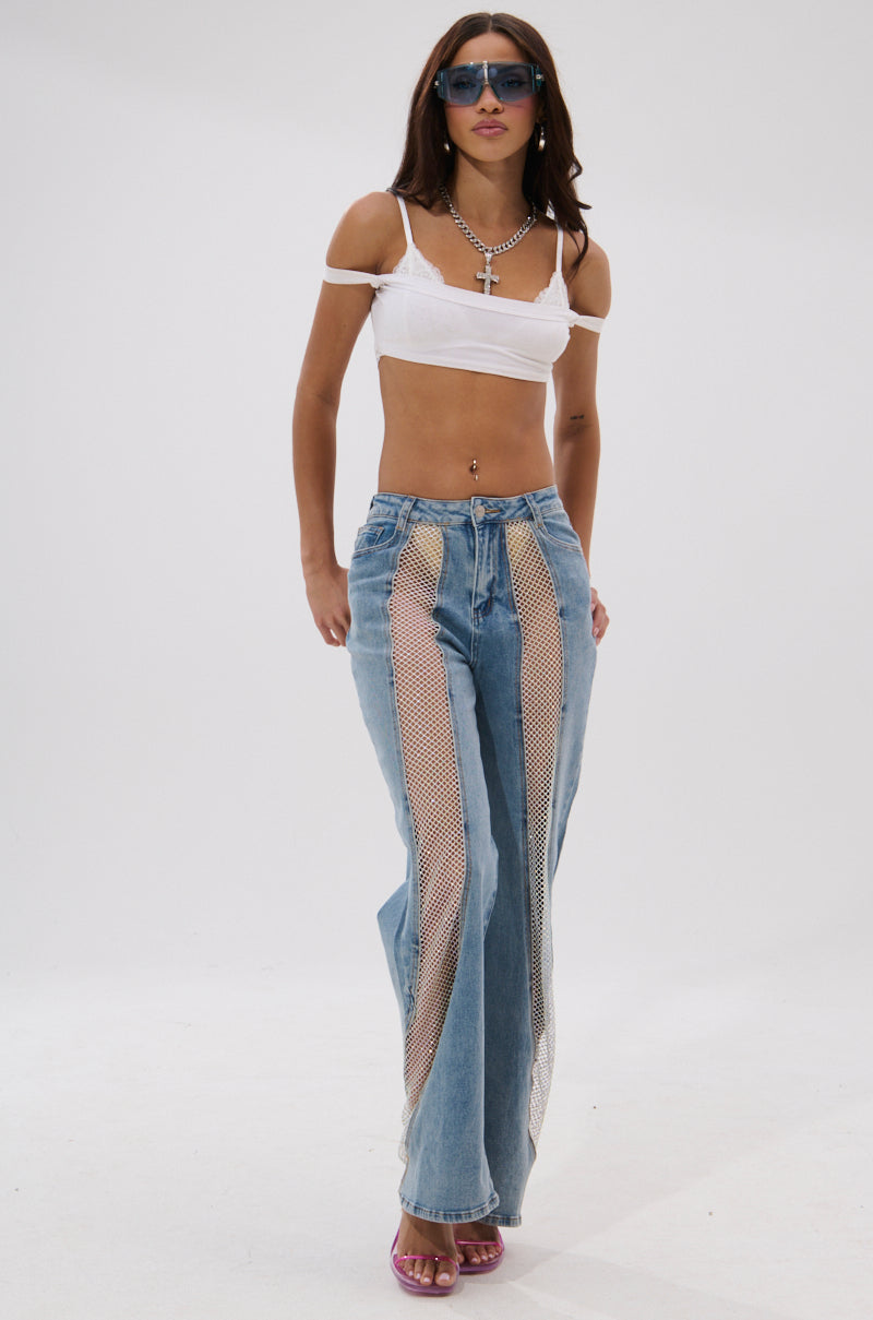 A woman wearing a white crop top, BLING BLING RELAXED DENIM jeans with sheer mesh panels, pink heels, layered necklaces, and blue sunglasses stands against a plain white background. 6