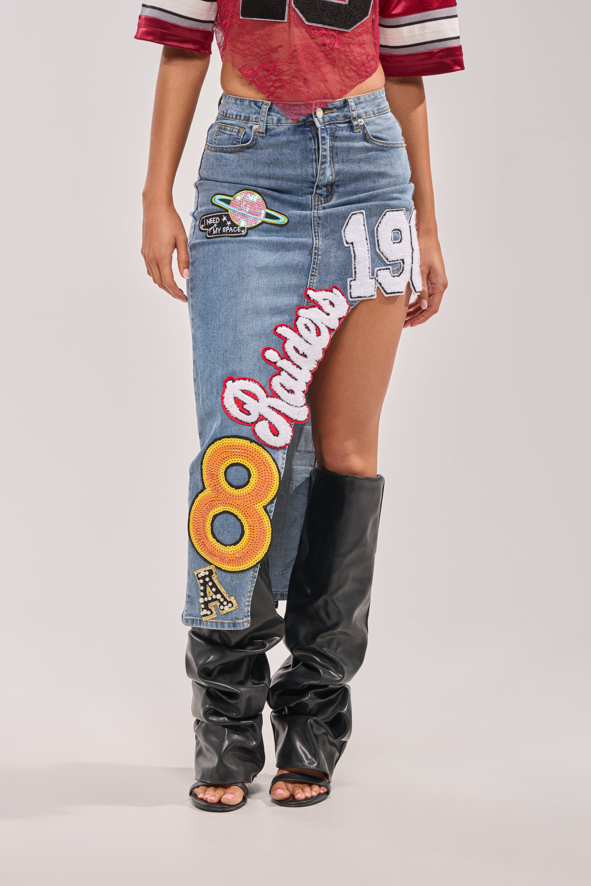A person wears the RAIDERS DENIM MAXI SKIRT featuring colorful patches—number 8, 19, "Riders," and a planet—with slouchy black boots and a cropped, sporty red shirt. 0