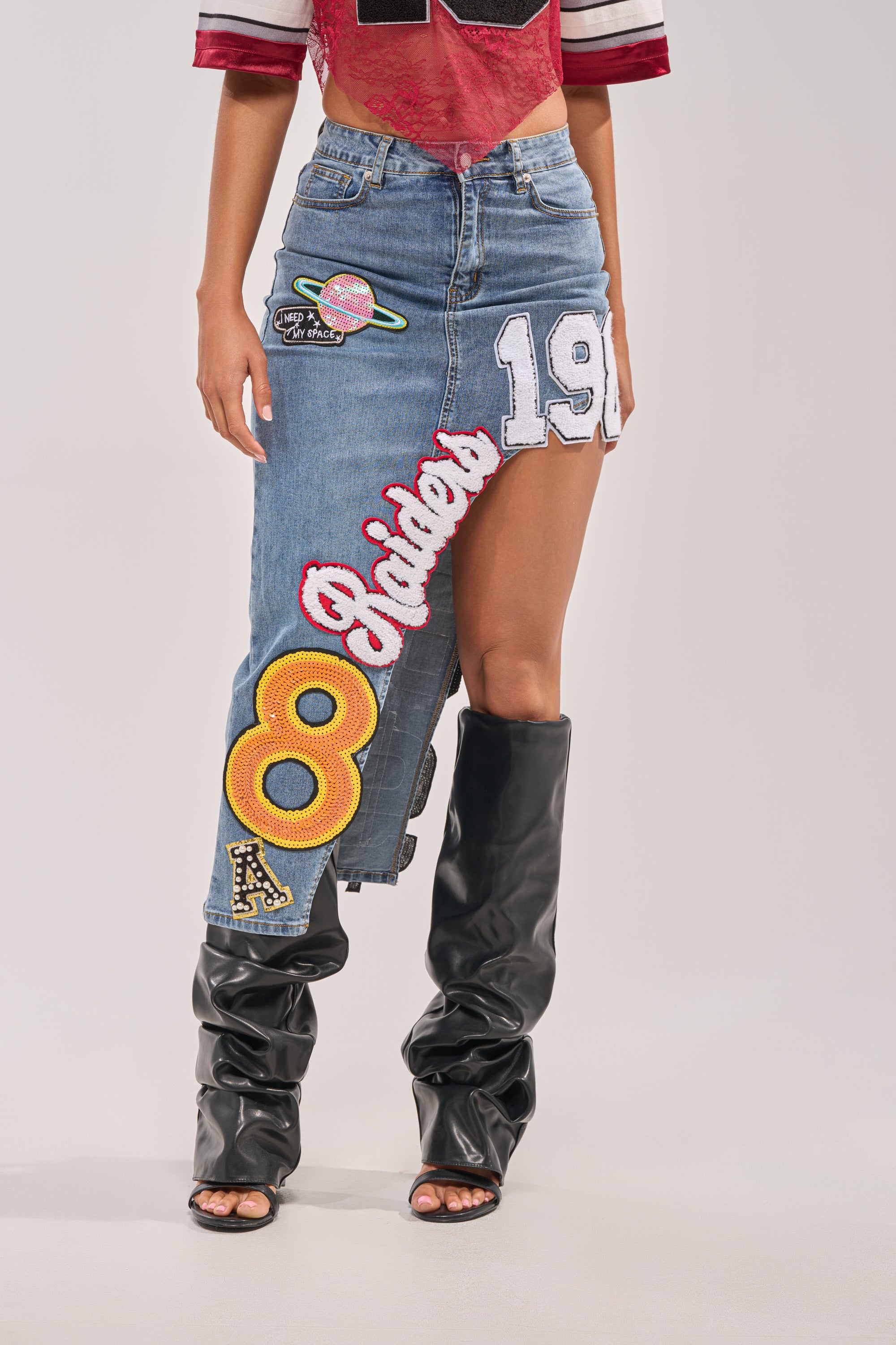 A person wears a red crop top, the RAIDERS DENIM MAXI SKIRT with bold varsity numbers and planet patches, and black slouchy knee-high boots—a perfect blend of game day streetwear and standout style. 1