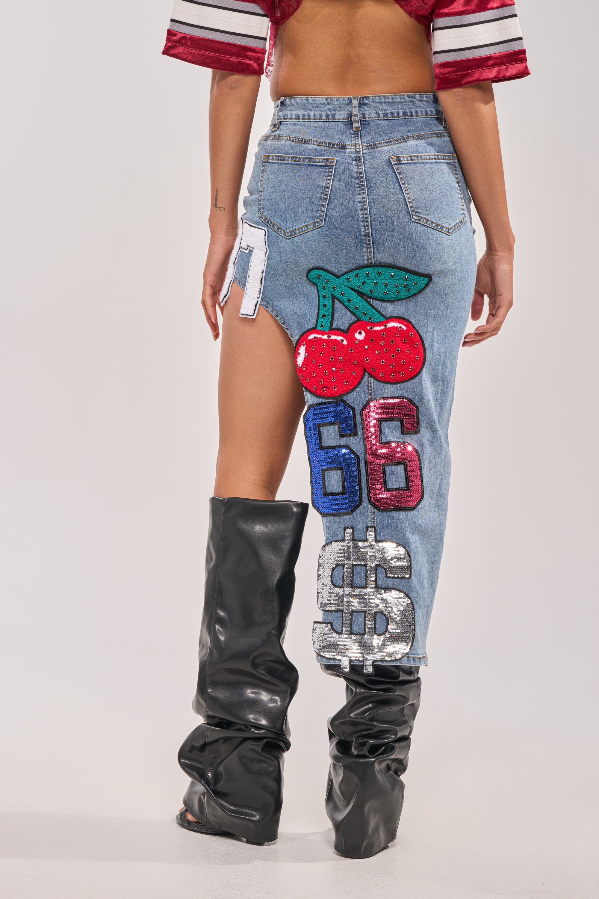 A person sporting the RAIDERS DENIM MAXI SKIRT with bold patches—cherries, “66”, a dollar sign, and a white patch—styled with oversized black slouchy boots and a red cropped top for cool game day streetwear vibes. 3
