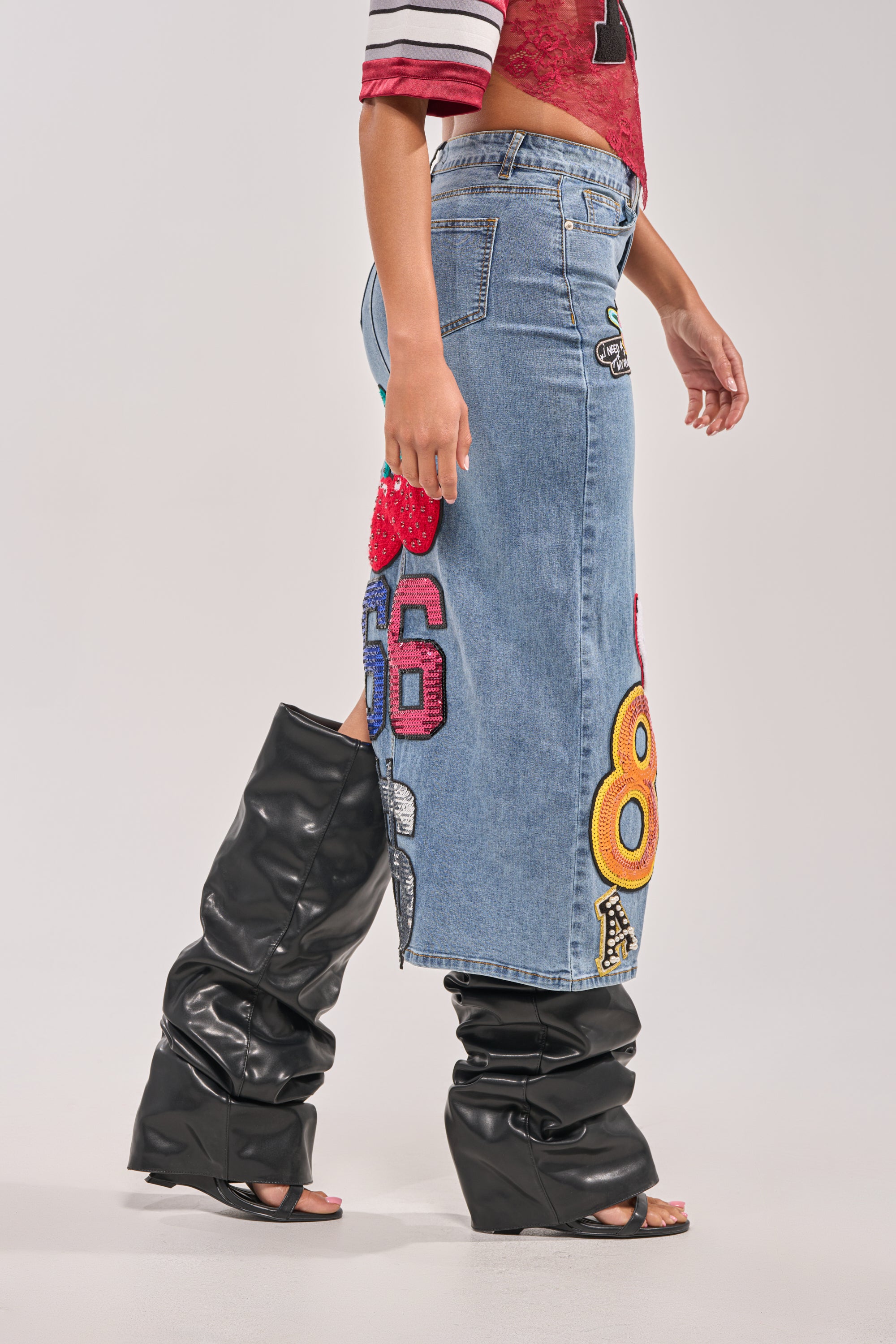 A person wearing the RAIDERS DENIM MAXI SKIRT with colorful number patches, a red and white cropped top, and black slouchy heeled boots is photographed from the side against a plain background for game day streetwear inspo. 4
