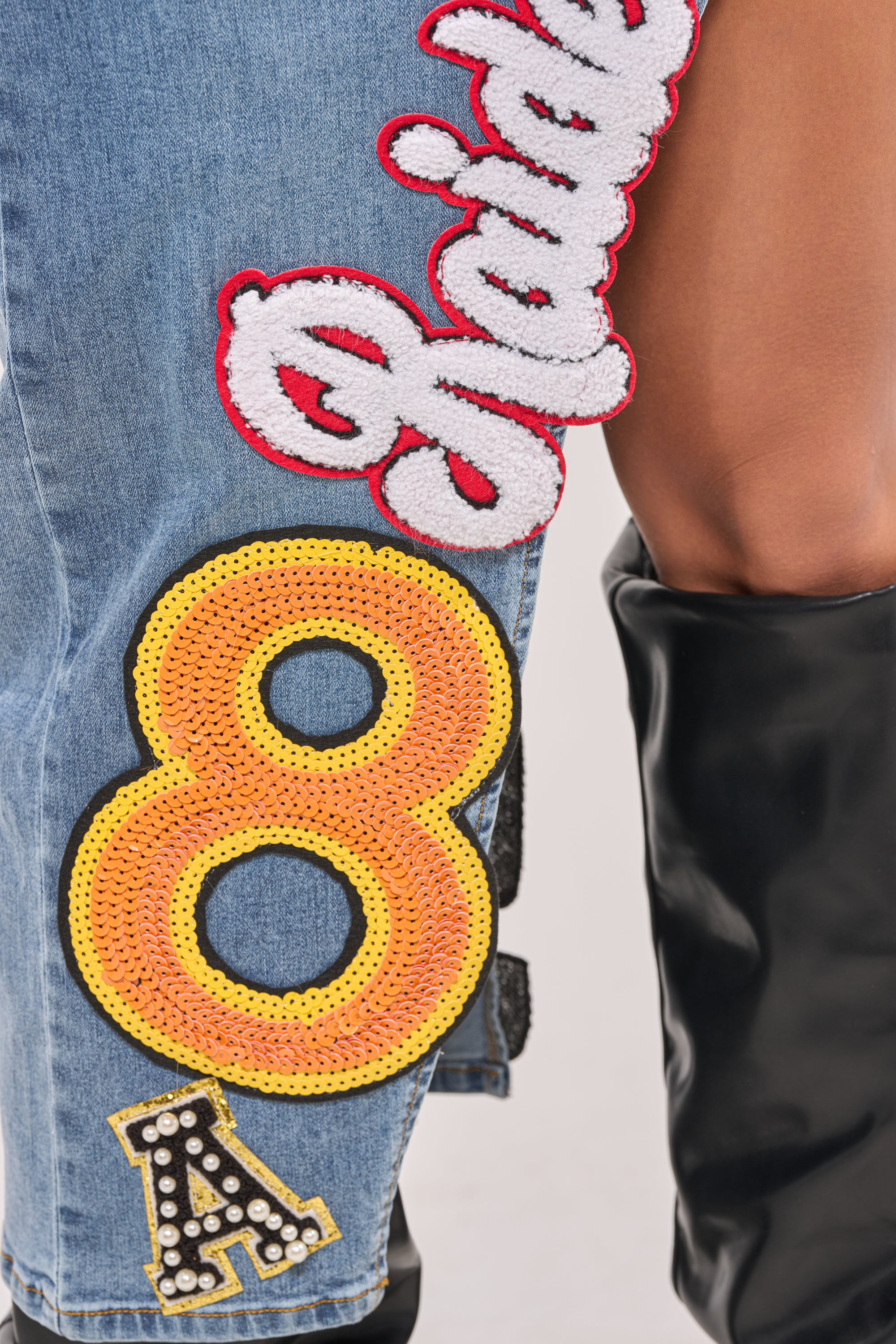 Close-up of a person wearing the RAIDERS DENIM MAXI SKIRT, featuring blue denim with colorful sequin patches, varsity “Raiders” details, a yellow number 8, and a black and gold letter A, paired with black boots. 6