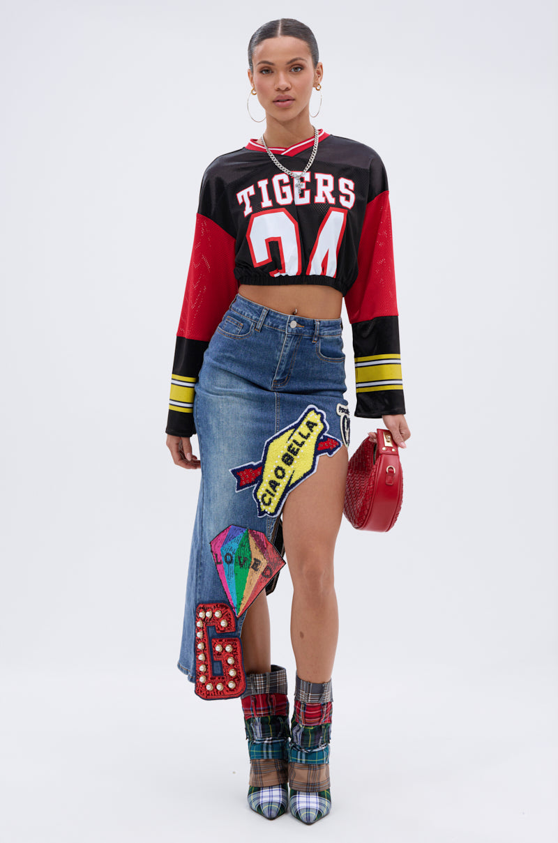 A woman poses against a white background wearing a cropped "Tigers 24" football jersey, the VARSITY SQUAD DENIM GRAPHIC MIDI SKIRT with colorful patches, plaid boots, hoop earrings, and holding a red handbag. 0