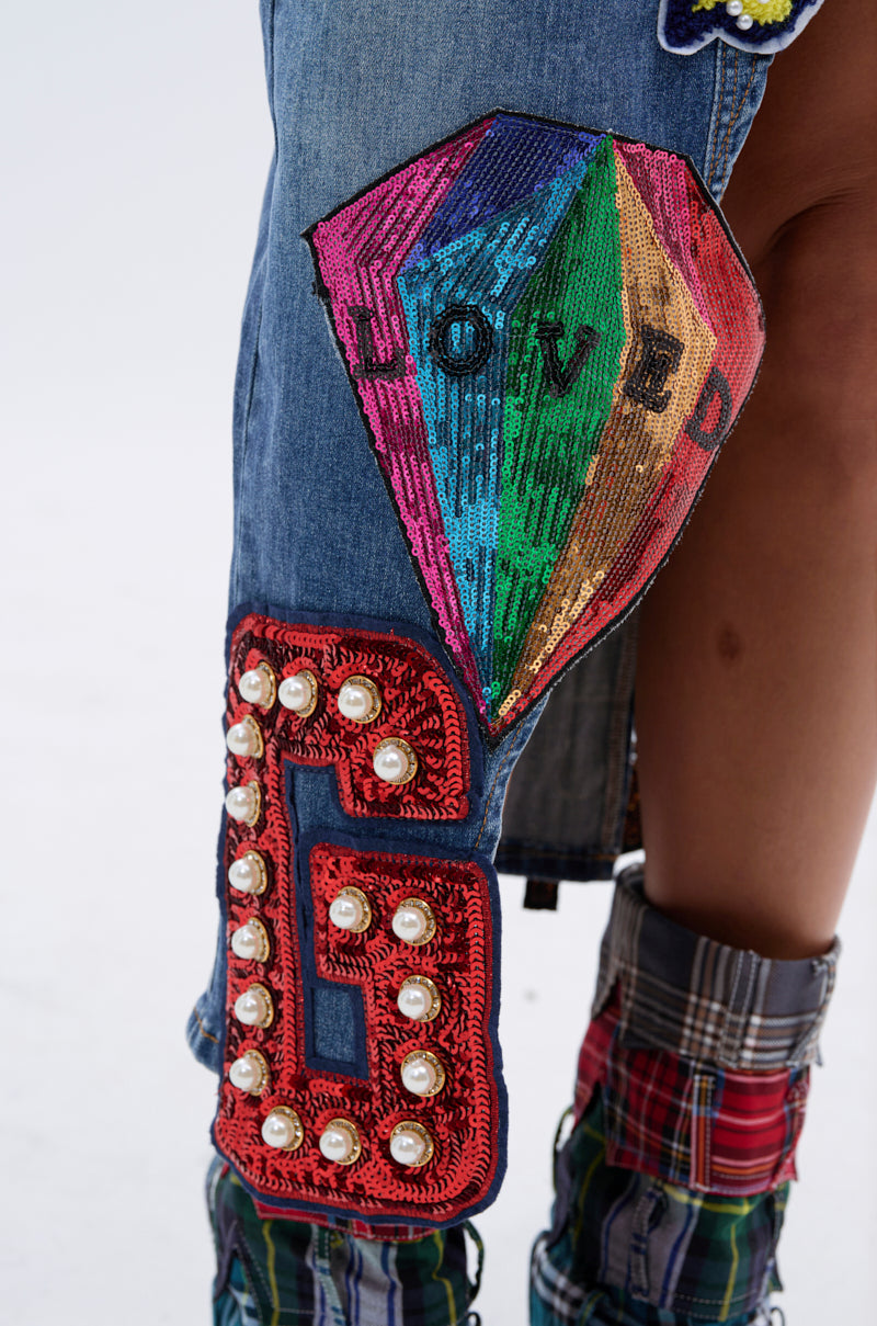 Close-up of the VARSITY SQUAD DENIM GRAPHIC MIDI SKIRT with a colorful sequin "LOVED" diamond patch and a red varsity-style "G" patch embellished with pearls. Plaid fabric and boots are also partially visible. 9