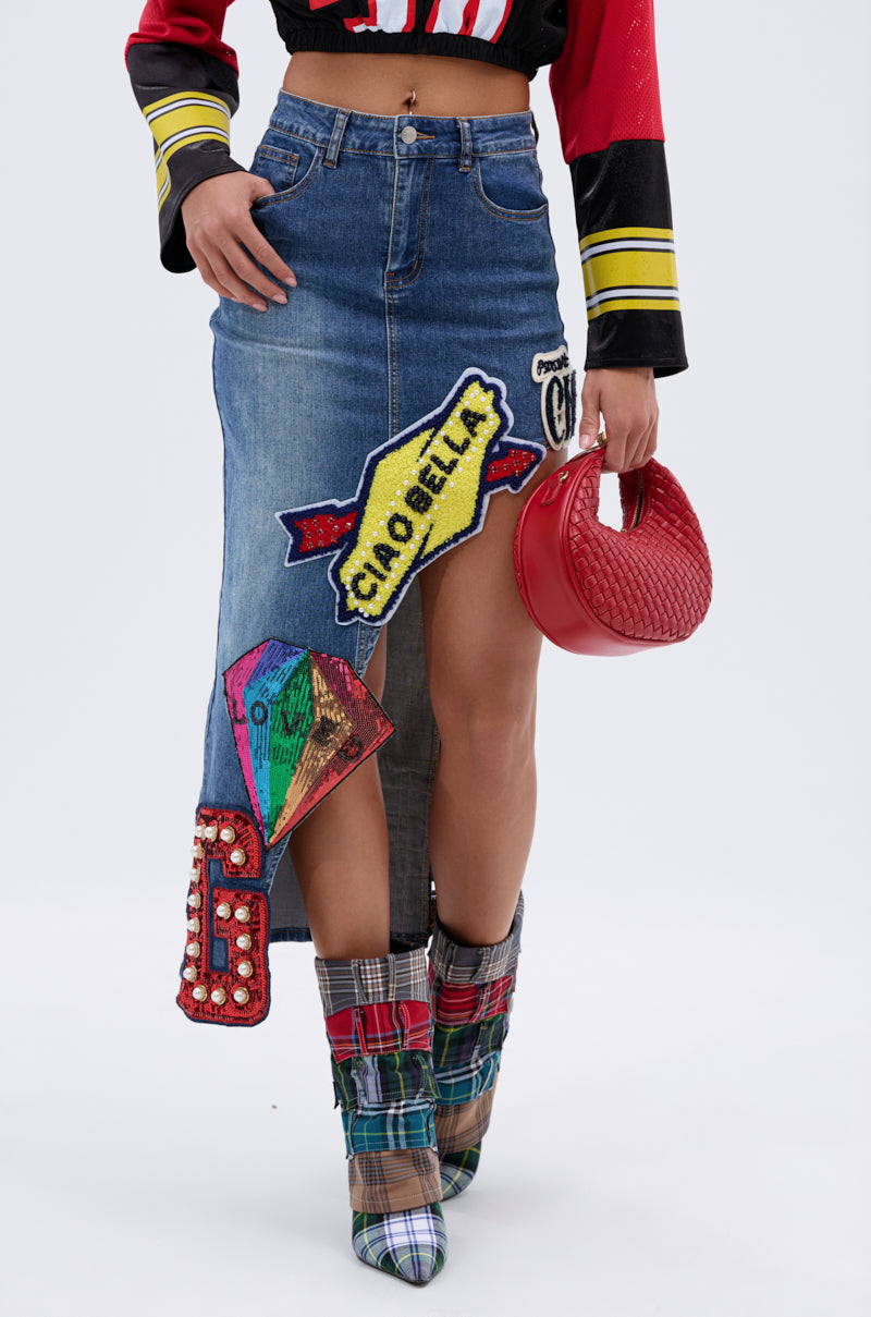 A person wearing the VARSITY SQUAD DENIM GRAPHIC MIDI SKIRT with colorful patches, a striped crop top in red, yellow, and black, plaid high-heel boots, and holding a small red textured handbag. 1