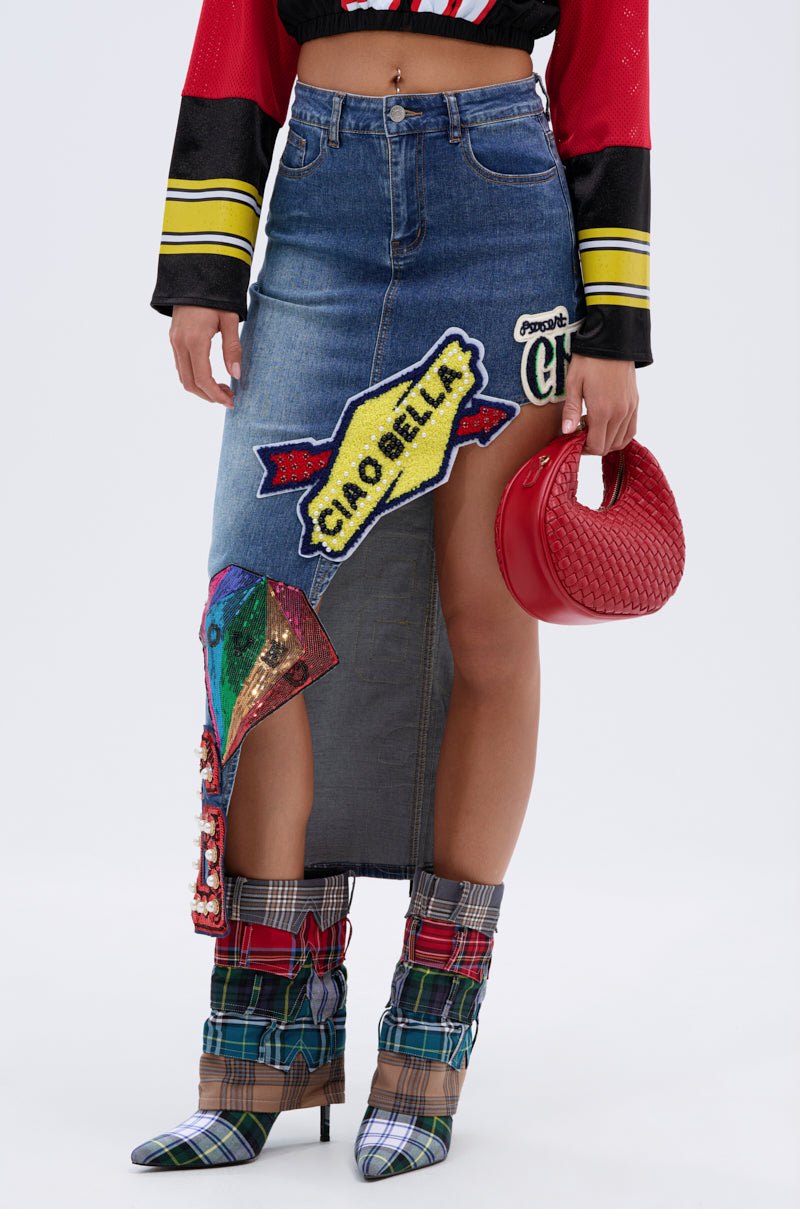 A person wearing the VARSITY SQUAD DENIM GRAPHIC MIDI SKIRT with colorful patches, a red and black top, plaid high-heeled boots, and holding a small red woven handbag. Their face is not visible. 2