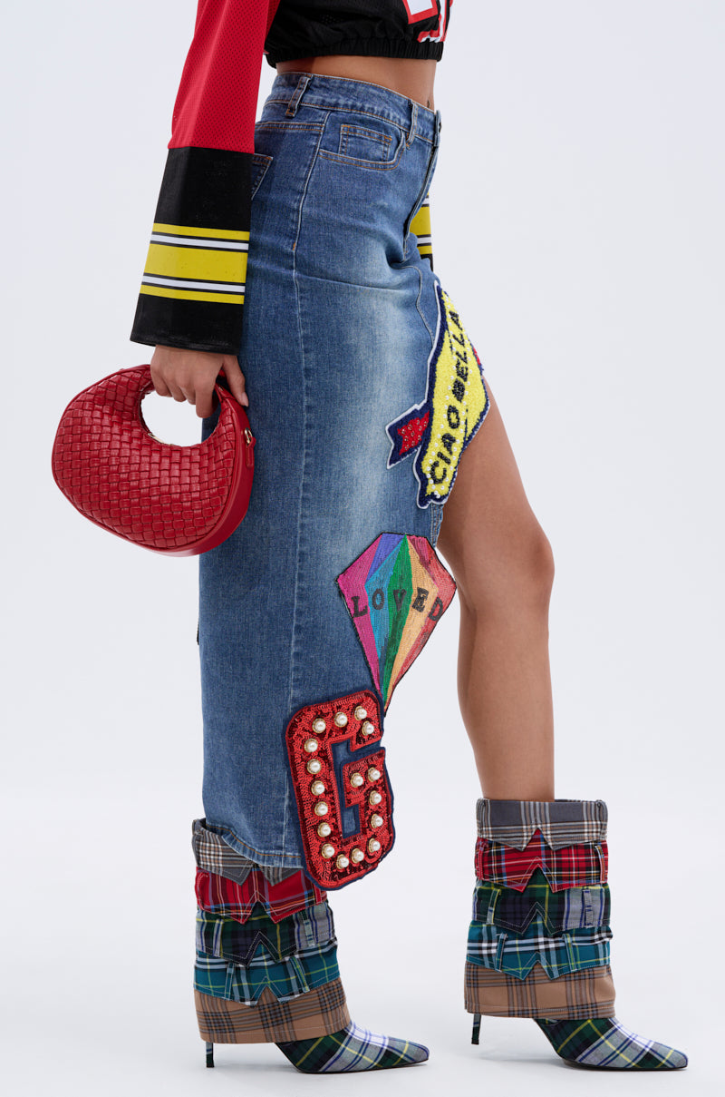 Wearing the VARSITY SQUAD DENIM GRAPHIC MIDI SKIRT with bold “G” and “LOVE” designs, plaid heeled boots, a black and yellow long-sleeve top, and carrying a small red woven handbag. 3