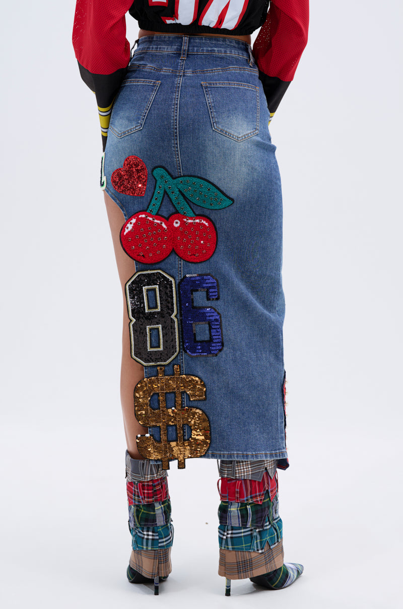 Someone wears the VARSITY SQUAD DENIM GRAPHIC MIDI SKIRT, detailed with colorful sequin patches—heart, cherries, “86,” and a dollar sign—styled with plaid leg warmers and open-toe heels. 5