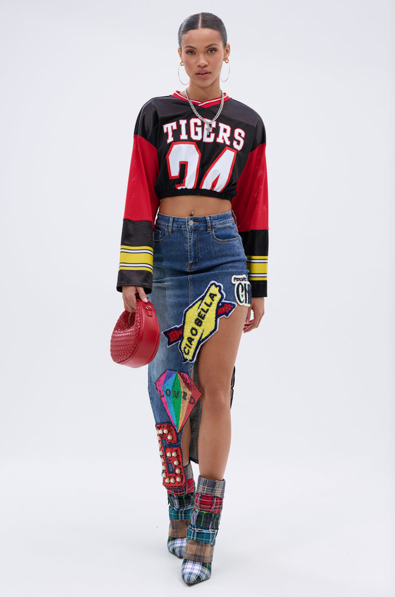 A woman wears a cropped black and red “Tigers 24” jersey, plaid heeled boots, hoop earrings, a red textured handbag, and the VARSITY SQUAD DENIM GRAPHIC MIDI SKIRT with colorful embroidered patches. She stands against a white background. 6